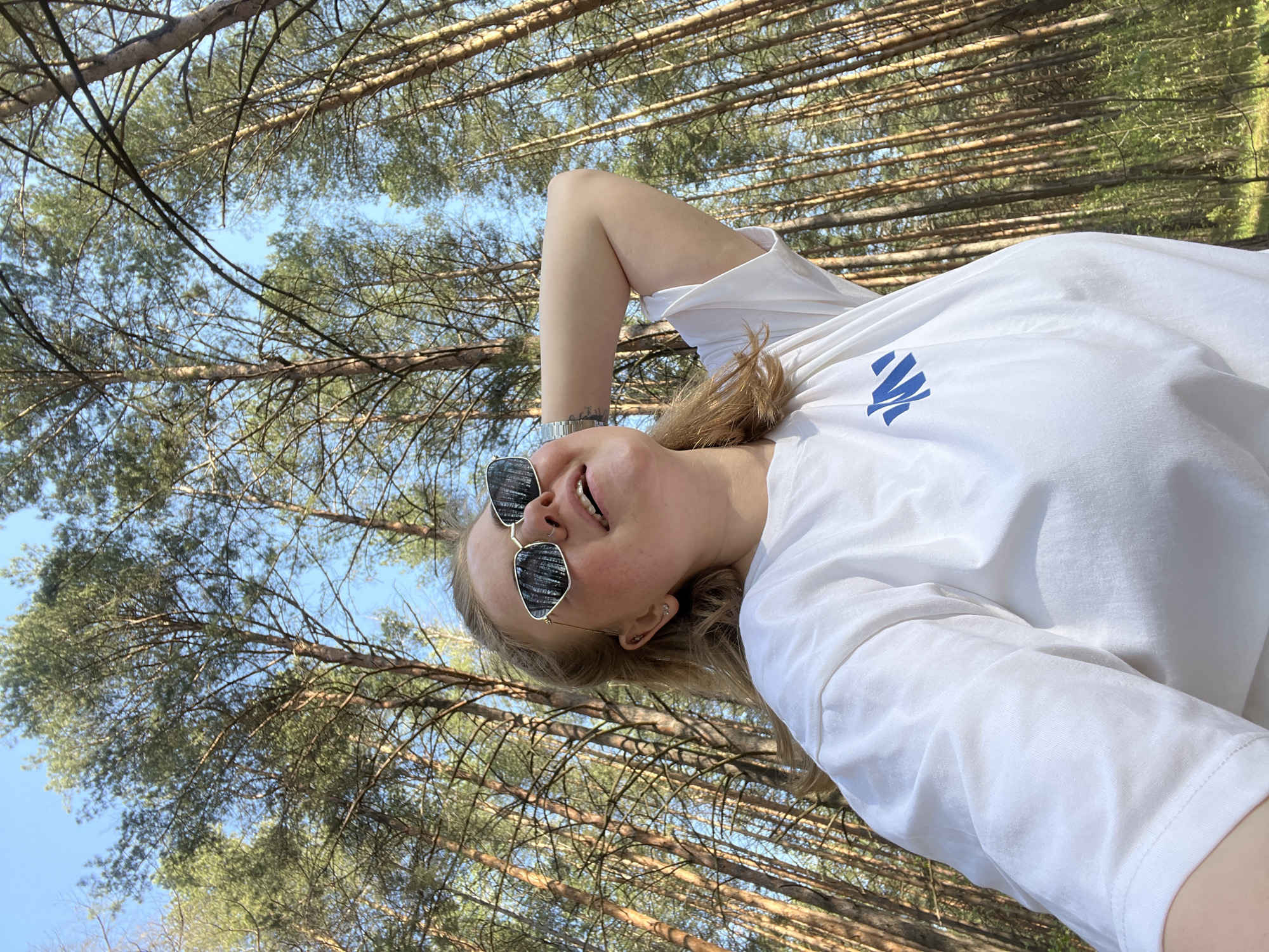 A young woman with blonde hair, wearing a white t-shirt and sunglasses, is lying on the ground in a forest. The trees are tall and thin, and the sky is visible through the branches. The woman is smiling and looking up at the camera. The location is likely a forest in the city of Chaikovsky, Russia.