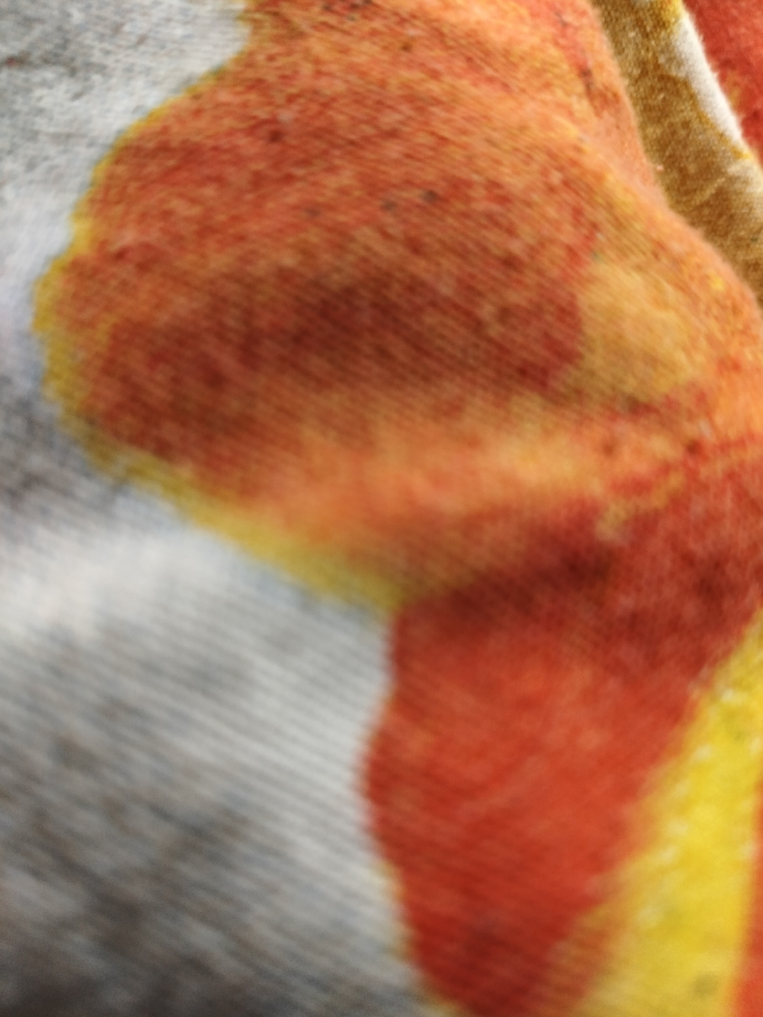 The image is a close-up, out-of-focus view of a textured fabric with a gradient of colors, primarily shades of orange, yellow, and gray. The fabric appears to be woven, with visible threads creating a ribbed texture. The colors blend into one another, suggesting a tie-dye or patterned design. There are no discernible objects, people, actions, or interactions visible in the image. The location in the city of Askino, Russia, is not discernible from the visual information provided in the image.