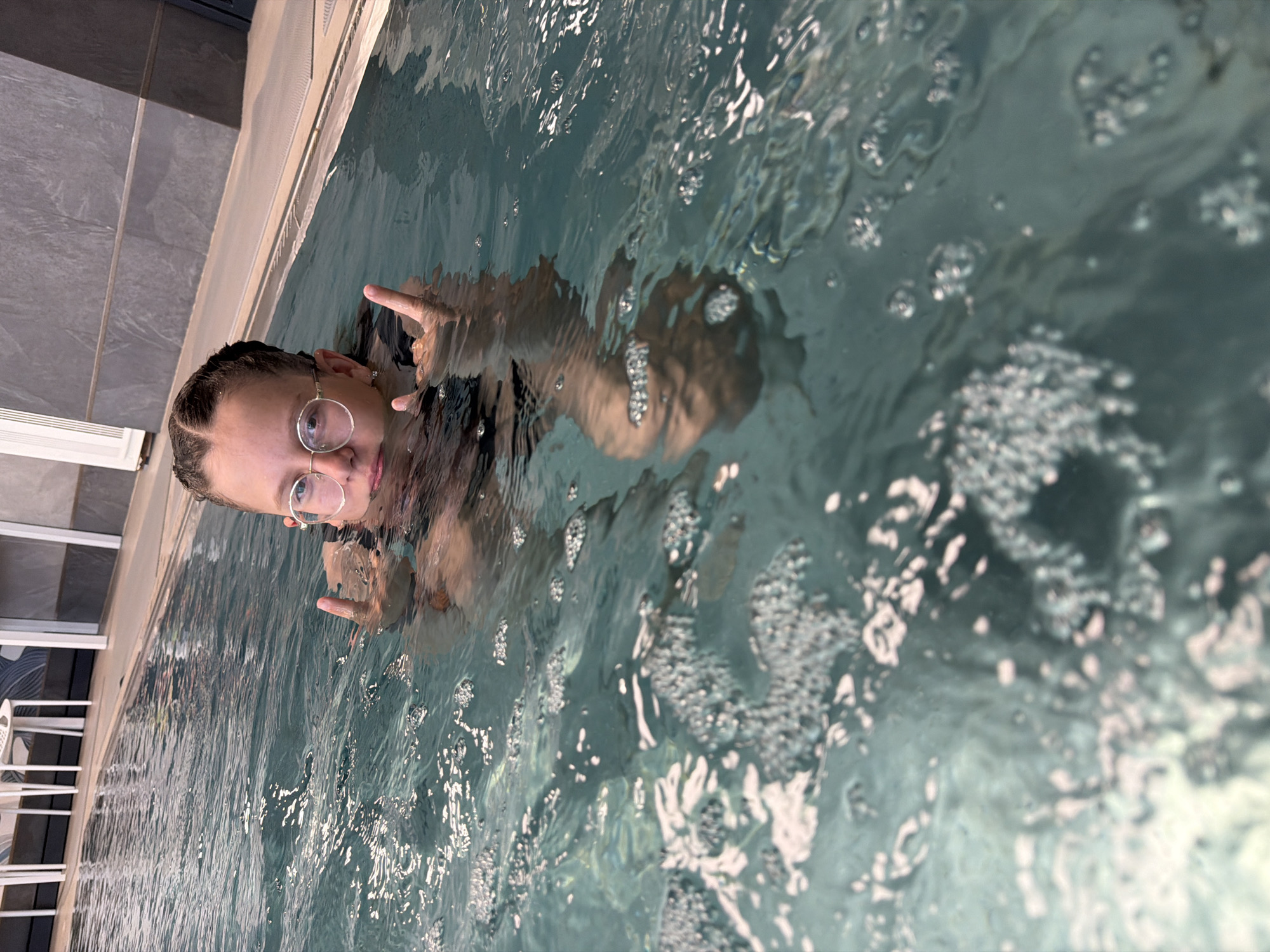 A person with braids and glasses is in a pool of water, facing the camera and making a hand gesture. The water is rippling and has bubbles. The edge of the pool is visible in the background, along with what appears to be a chair.