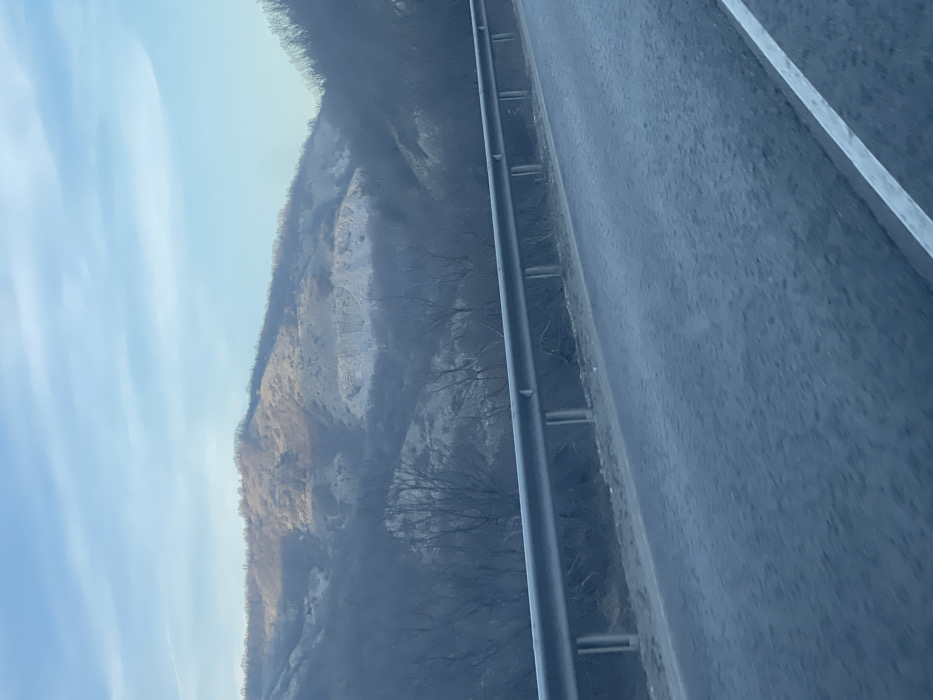 The image depicts a rural mountainous or hilly landscape, observed from a low angle, likely from a moving vehicle, in the vicinity of Paidushko, Bulgaria. A two-lane asphalt road with a visible white solid line marking the right edge occupies the lower foreground. A galvanized steel guardrail runs parallel to the left side of the road, separating it from the adjacent terrain. Beyond the guardrail, the immediate hillside is covered with dark, bare deciduous trees and some visible ground. In the midground and background, multiple layers of hills are visible, predominantly covered in dormant, leafless vegetation. Patches of lighter-colored, possibly rocky or cleared, terrain are interspersed across the hillsides. The distant hills show areas illuminated by sunlight, contrasting with the shadowed lower slopes and foreground. The sky is light blue with scattered, thin stratiform clouds. No people, animals, or specific actions are discernible. The overall lighting suggests late afternoon or early morning conditions in a cold season.