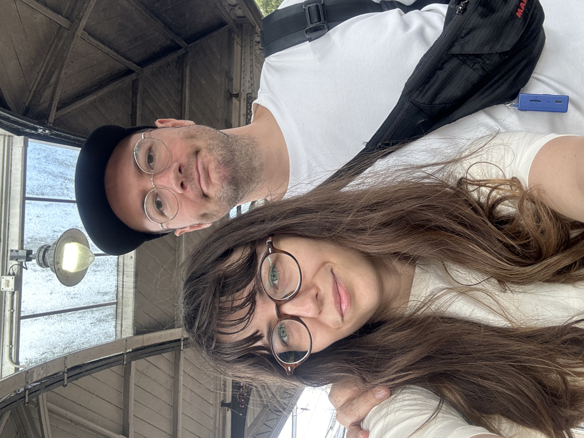The image shows a close-up selfie of a couple in an outdoor urban setting. The woman is in the foreground, wearing glasses and a light-colored top, with her brown hair falling around her shoulders. The man is behind her, wearing a black baseball cap and glasses, and a white t-shirt. He has his arm around the woman. He is wearing a black fanny pack across his chest, with a small blue electronic device attached to it. The background shows the roof of a structure with a glass-paneled ceiling and wooden beams, illuminated by an industrial-style light fixture. The overall lighting is soft and diffused, suggesting an overcast day. The location is identified as Norwich, United Kingdom, likely a covered walkway or waiting area, possibly at a train station or market given the structure's design. The focus is on the couple's faces, with a slight blur on the background elements.