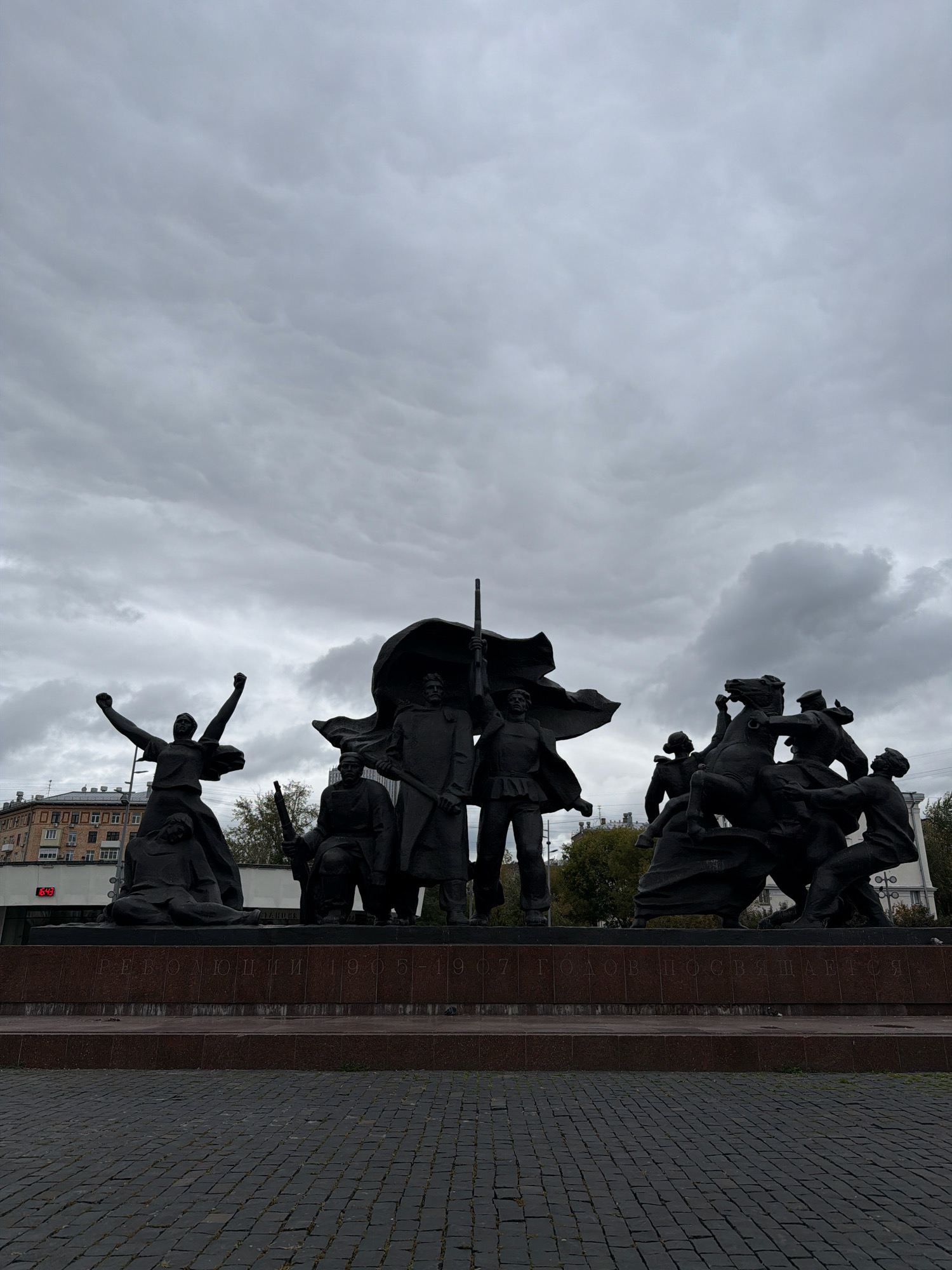 The image depicts a monumental bronze sculpture group situated in Moscow, Russia, in front of buildings under a cloudy sky. The sculpture, located on a granite base, features several figures. In the foreground left, a female figure with arms raised in a gesture of triumph stands over a fallen figure. To the right of center, a group of figures is depicted in dynamic action, with one figure leading a horse ridden by another figure. Behind these, a man stands holding a rifle and a flag. A partially visible inscription on the granite base reads "РЕВОЛЮЦИИ 1905-1907 ГОДОВ ПОСВЯЩАЕТСЯ", translating to "Dedicated to the Revolutions of 1905-1907". The ground in front of the monument is paved with dark cobblestones. The overall atmosphere is somber, likely due to the overcast weather and the subject matter of the sculpture.