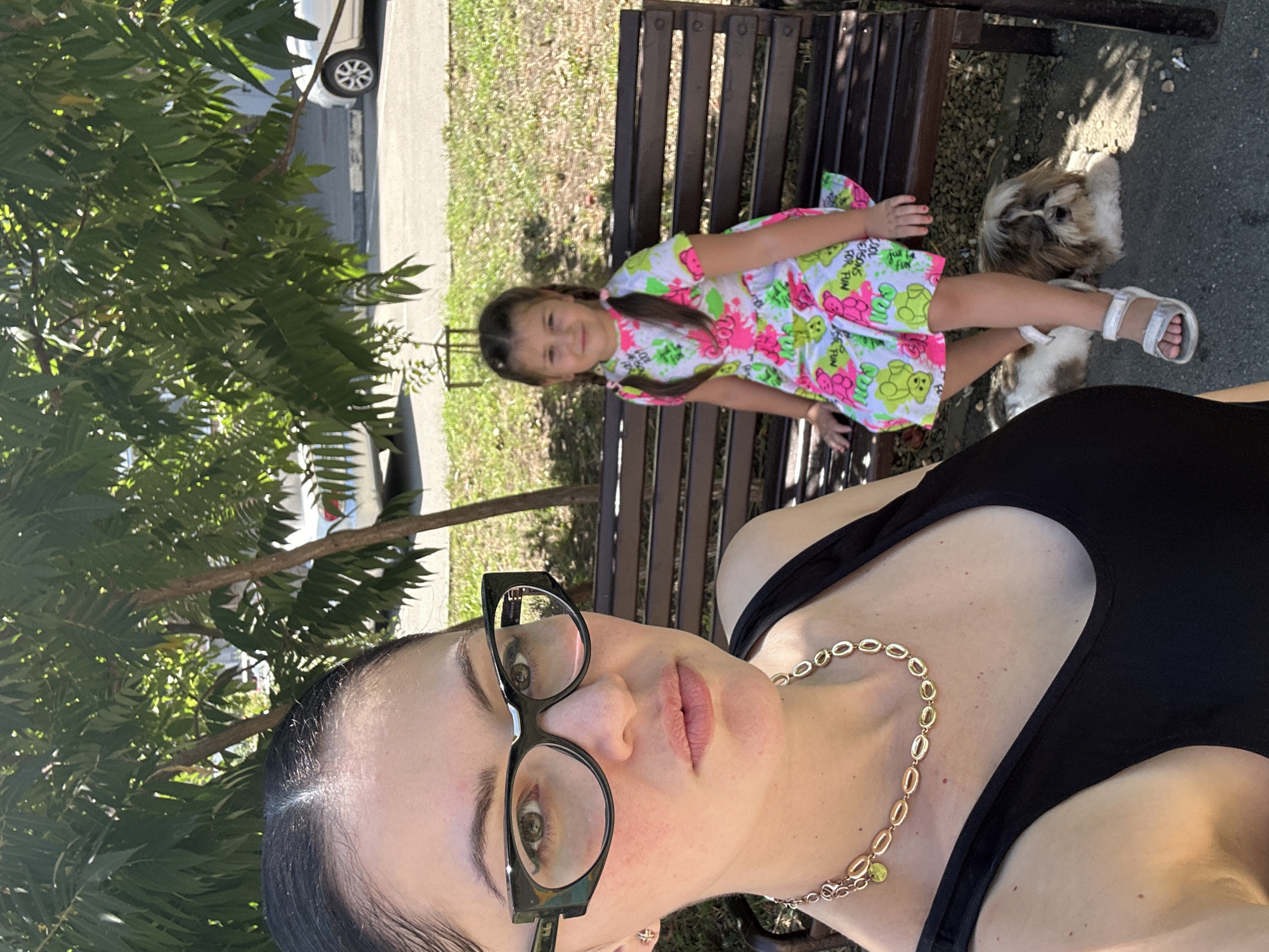 In an outdoor, sunny setting, a woman is taking a selfie. She is wearing a black tank top and a gold necklace with oval-shaped pendants. She has dark hair pulled back and is wearing large-rimmed black glasses. To her right, a young girl in a colorful dress with bears and neon accents sits on a wooden bench. The girl has her hair in pigtails and is wearing white sandals. A small shih tzu dog lies on the ground next to the girl. Behind them, a wooden bench is visible, and beyond that, a paved area with a parked white car. The environment includes green trees and grass. The location is described as Unknown, Unknown.