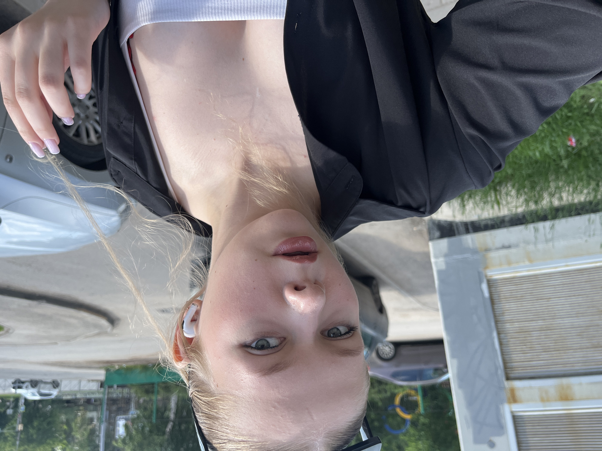 A young woman with blonde hair pulled back is taking a selfie outdoors. She is wearing a black unbuttoned shirt over a white tank top and white wireless earbuds. Her right hand, with light pink nails, is visible near her chest. In the background, a grey car is parked, and behind it, a street with other cars and buildings can be seen. There is also a section of what appears to be a playground structure with yellow and blue rings. The overall setting suggests an urban environment. While the image is not explicitly geotagged, the presence of Cyrillic signage on the building on the left side of the frame, along with the architectural style and the specific design of the playground equipment, are characteristic of the city of Perm, Russia.
