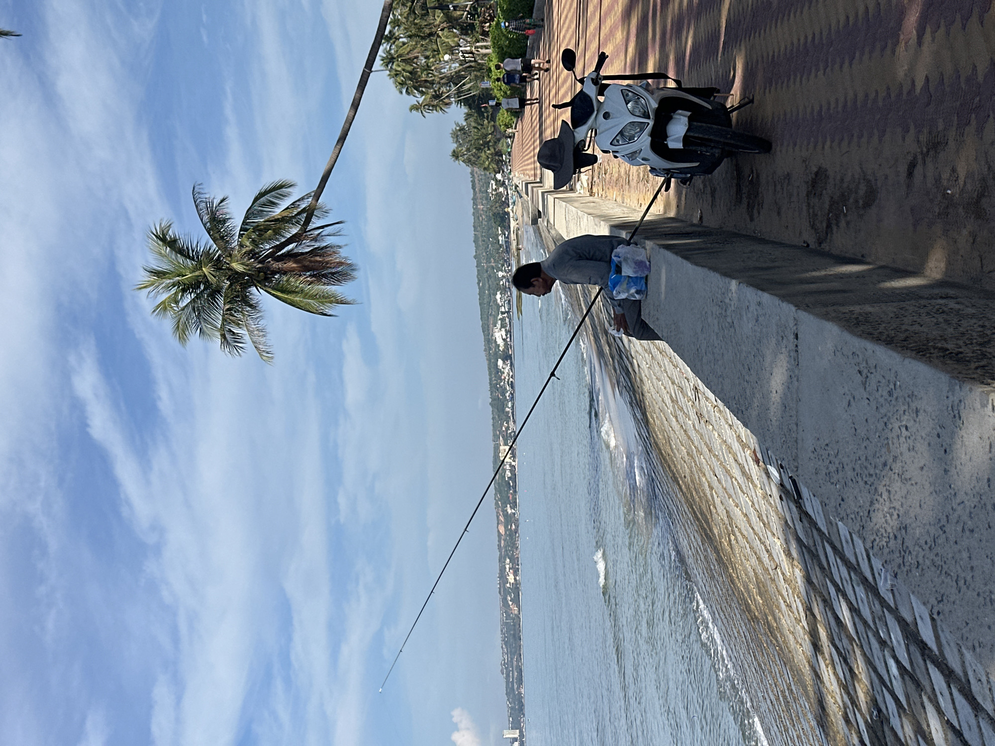 A man is fishing by the sea on a paved walkway in Mui Ne, Vietnam. He is sitting on a low concrete wall, holding a fishing rod that extends into the water. Beside him, on the walkway, are a white motorcycle and a hat. A coconut palm tree leans over the walkway, its fronds casting shadows. In the distance, buildings and lush vegetation line the coast. Other people can be seen walking along the walkway further down. The sea is calm with gentle waves lapping against the shore. The sky is blue with scattered white clouds.