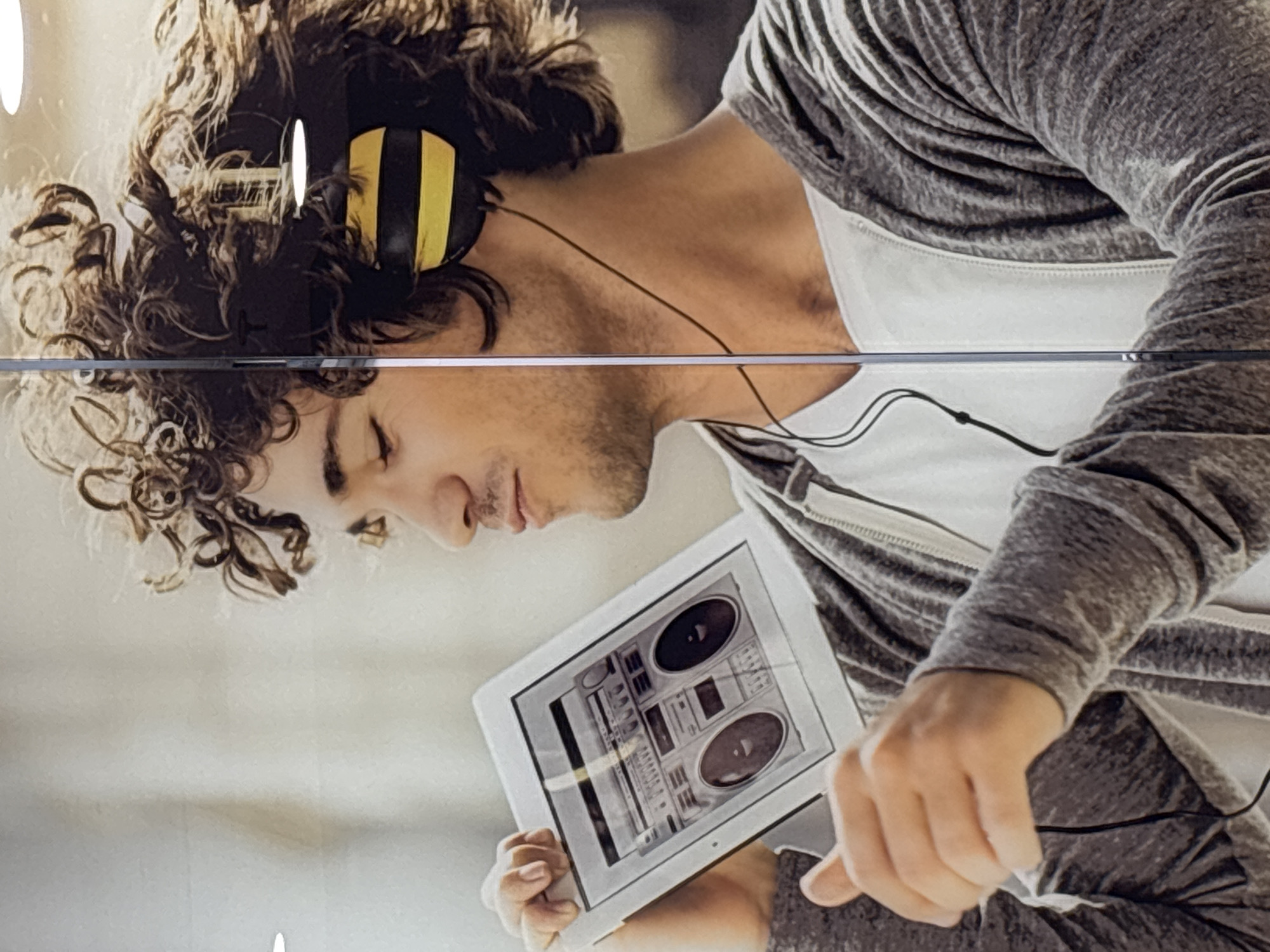 A close-up, indoor shot shows a person with curly brown hair and wearing yellow and black headphones. They are listening to music on a tablet which has an image of a boombox on the screen. The person is wearing a white zip up jacket with a gray knit sweatshirt over it. The person's eyes are closed. The scene is indoors and well-lit. The location is unknown.