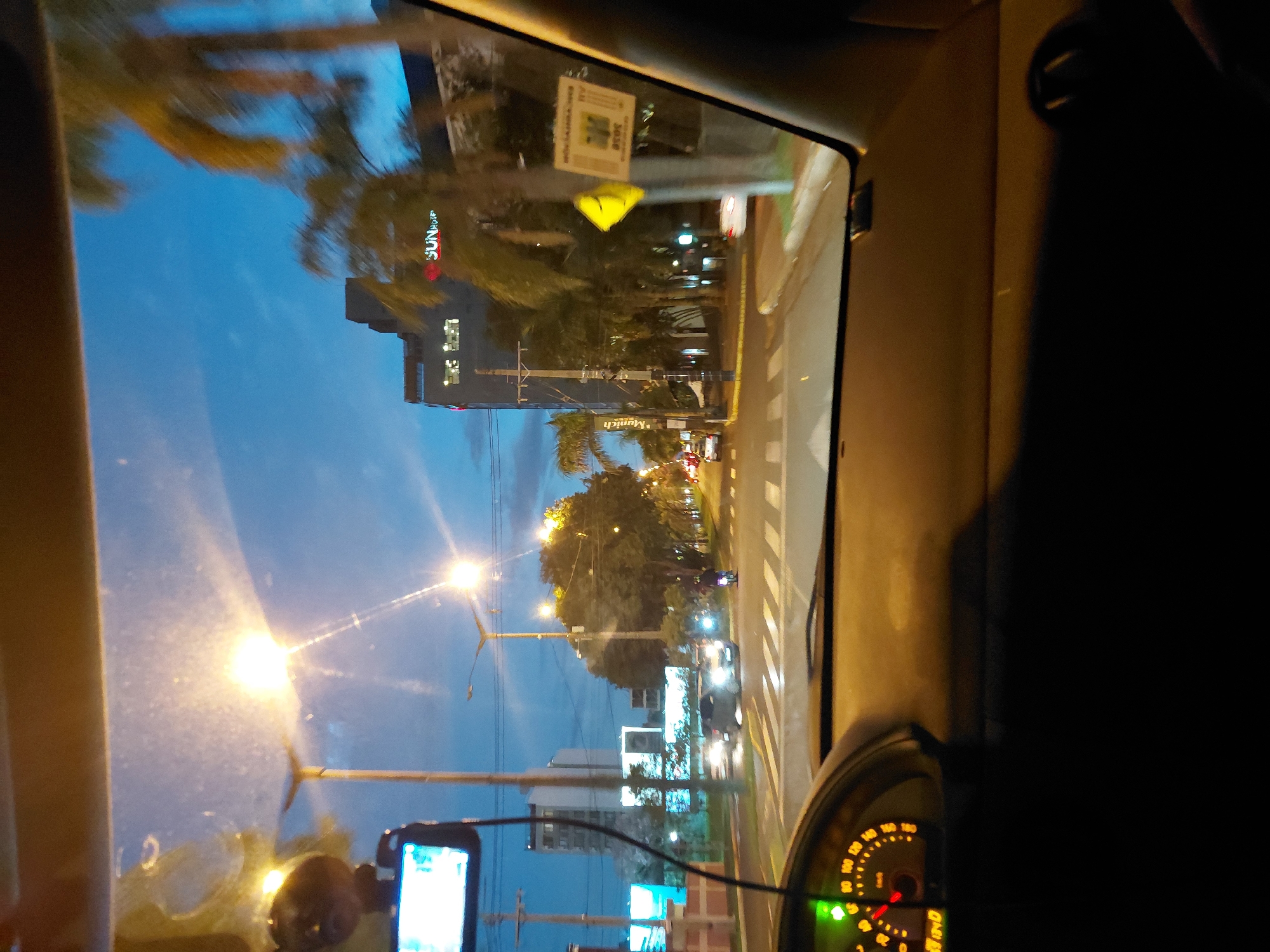 The image, taken from inside a vehicle, captures a view of a city street at dusk in Encarnación, Paraguay. The foreground features the dashboard of the car, with a lit speedometer indicating a speed of approximately 20 km/h. The windshield reveals a street scene with streetlights illuminating the road and surrounding buildings.
Several vehicles are visible on the road, including cars and a motorcycle. Pedestrian crossings are marked on the asphalt. Buildings line the street, some with illuminated windows, and palm trees are interspersed throughout the landscape, consistent with a tropical or subtropical urban environment. A prominent building with "SUN" logo is visible in the background, along with other multi-story structures. The sky is a deep blue, transitioning into dusk, with some clouds present. The overall atmosphere suggests a typical evening commute or drive through a city.