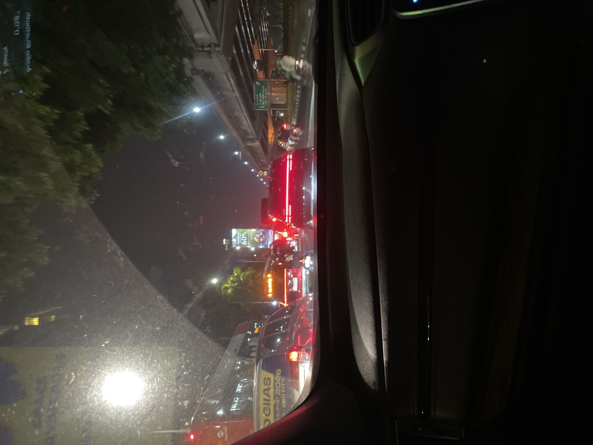 The image was taken at night from inside a vehicle in heavy traffic on a city street in South Jakarta, Indonesia. The perspective is looking through the windshield, which is speckled with raindrops. A red bus is visible on the left, partially obscured. Several cars are lined up on the road, with their brake lights illuminated. One car in the foreground has a yellow sticker on its rear window that reads "GIIAS" and "MALAYSIA 2026". A digital billboard displaying "60%" is also visible. On the right side of the road, an elevated highway structure is present, and there are streetlights and trees lining the road. Several motorcycles are also navigating through the traffic. The scene depicts a typical urban traffic jam at night.