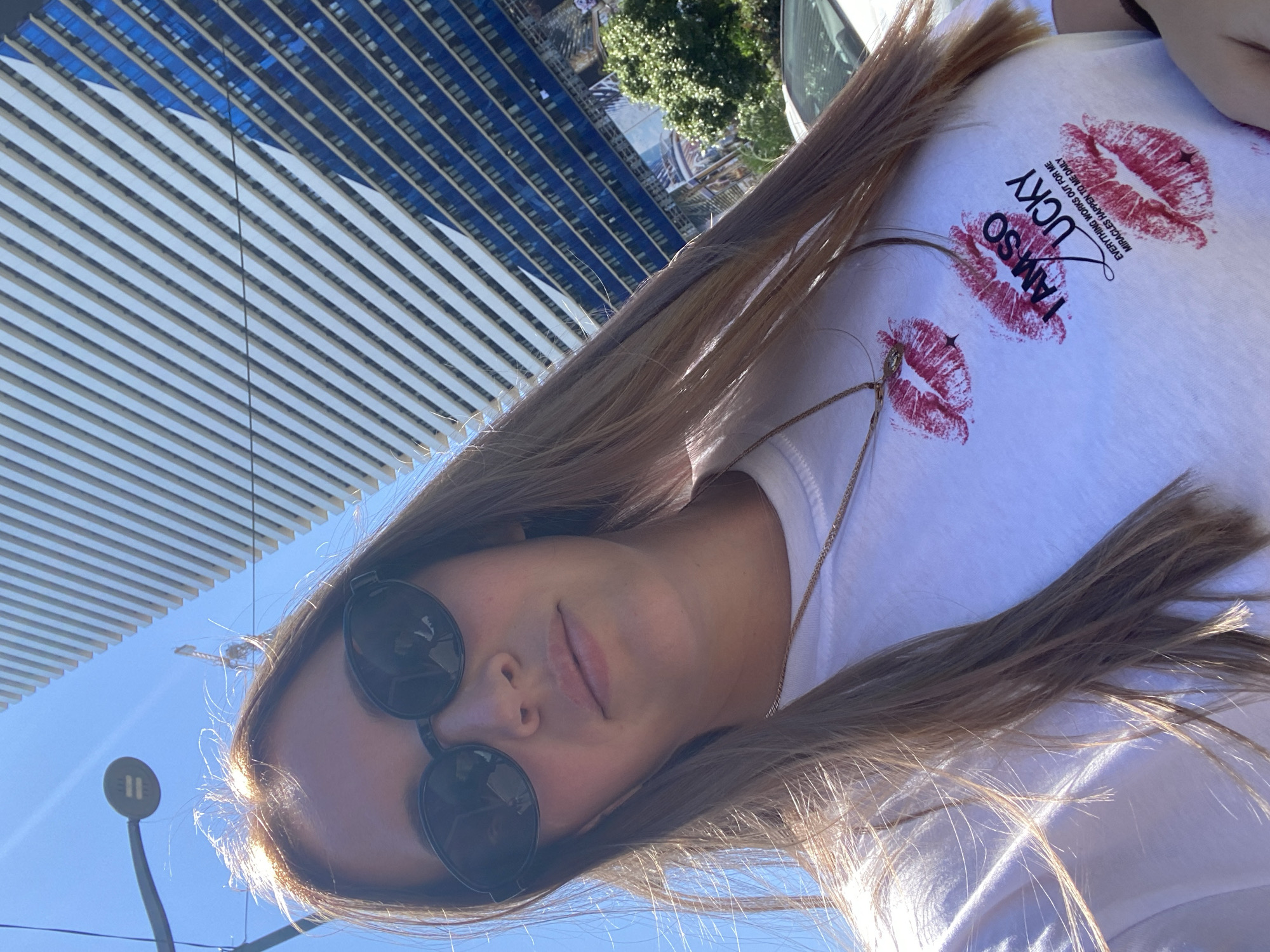 A young woman, wearing sunglasses and a white t-shirt with red lipstick kiss imprints and text that reads "I AM SO LUCKY EVERYTHING WORKS OUT FOR ME MIRACLES HAPPEN TO ME DAILY", is captured in a selfie. She is outdoors, with a clear blue sky above. In the background, a tall modern building with a blue facade and white vertical elements dominates the scene, likely in the city of Batumi, Georgia. A lamppost is visible to the left of the frame, and a portion of a white car is seen on the right. Sunlight casts shadows, highlighting the texture of her hair and clothing.