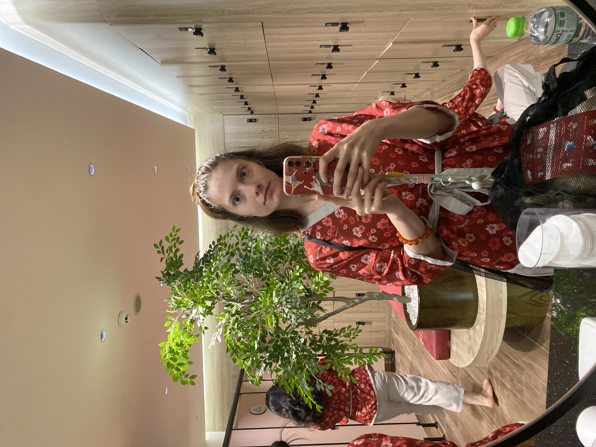 The image depicts a young woman taking a selfie in what appears to be a locker room with a spa-like ambiance. She is wearing a red robe with a floral pattern and has her hair tied up in a ponytail. In her right hand, she is holding a smartphone, capturing the reflection in a large mirror. The background reveals a row of wooden lockers, a tall artificial plant in a pot, and the reflective surface of a circular wooden bench. To the left, in the mirror's reflection, another person wearing a similar red robe is seen walking away, and further left, a portion of a changing cubicle is visible. On a counter in the foreground, a bottle of water and a container of cotton pads are placed. The overall setting suggests a wellness or leisure facility, potentially a spa or gym in Da Lat, Vietnam.