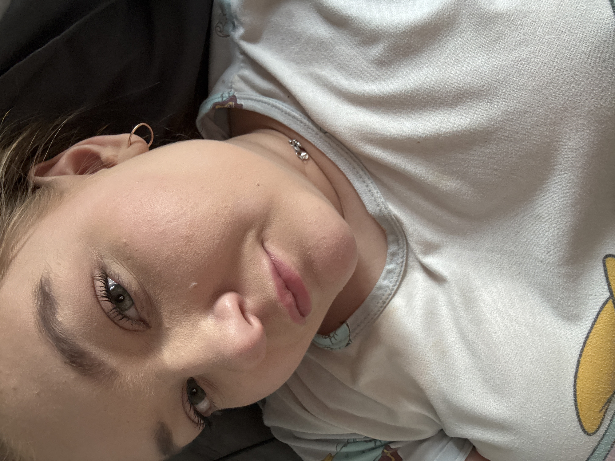 A close-up image depicts a young woman lying down, with a portion of her face and upper body visible. She has fair skin, light-colored eyes, and a small gold hoop earring in her left ear. She is wearing a light-colored top with a patterned trim around the neckline. A small silver pendant necklace is visible on her neck. The background includes a dark, undefined area that appears to be a pillow or bedding. There is no indication in the photograph that would give away the location as Chaikovsky, Russia.