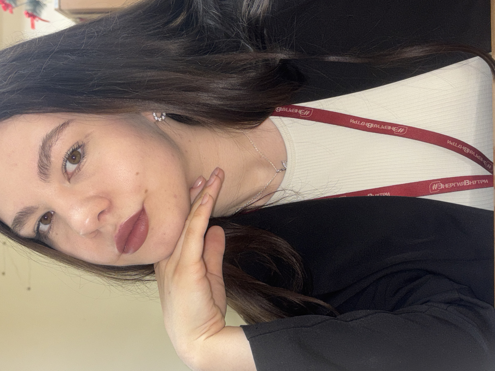 A young woman poses for a selfie. She has long brown hair, brown eyes, and is wearing dark lipstick. She is wearing a white ribbed top, a black blazer, and a silver necklace and earrings. Her right hand is touching her face. A red lanyard with Russian text "ЭНЕРГИЯВНУТРИ" (Energy Within) is visible around her neck. The background is blurred and suggests an indoor setting with light-colored walls. The location is potentially Perm, Russia, based on the text on the lanyard.