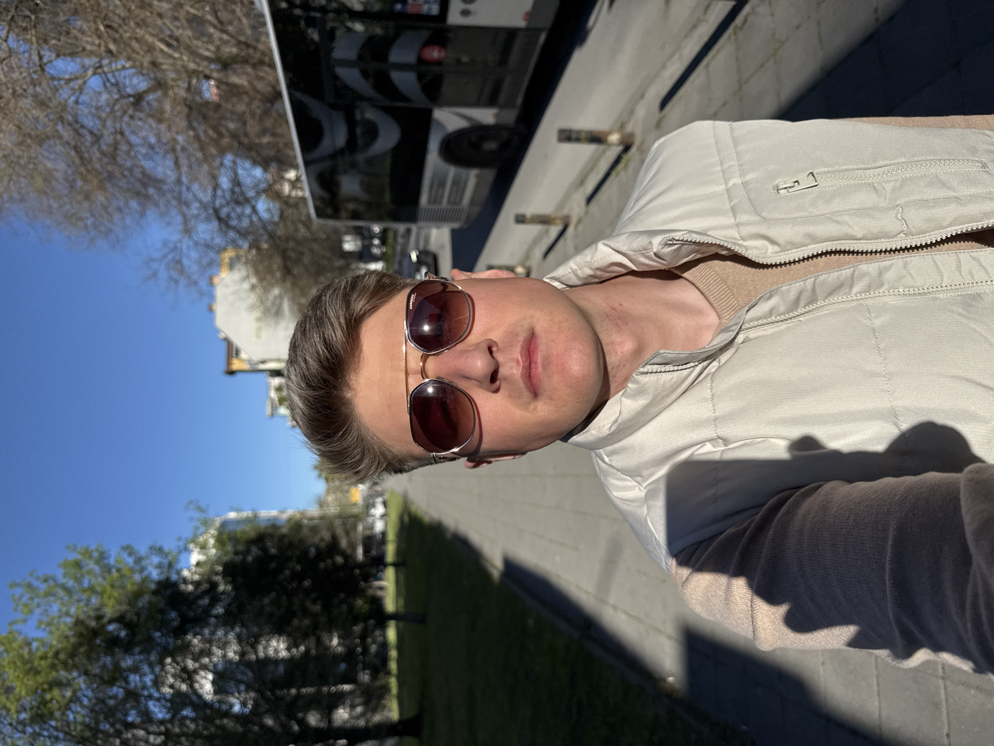 A man wearing sunglasses and a light-colored vest stands in the foreground. A bus is visible in the background, suggesting the location is near a bus stop or transit route. Trees line the street, partially obscuring buildings. The photograph was taken outdoors during the daytime under clear, sunny skies. The presence of buses and urban vegetation suggests an urban environment, but there is insufficient information to identify the location precisely in Plovdiv, Bulgaria.
