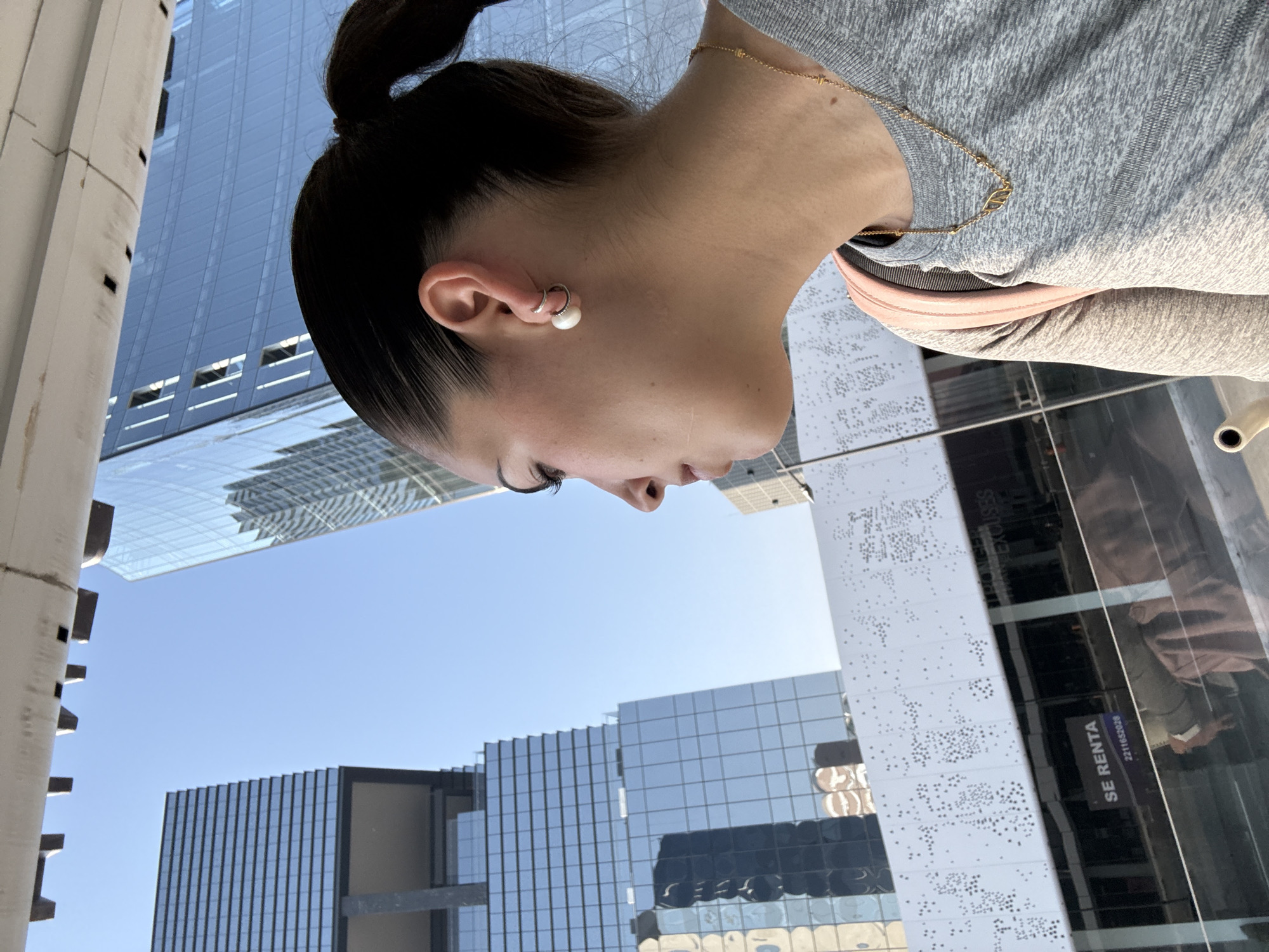 A woman with dark hair pulled back in a ponytail is the primary subject of an outdoor photograph. She is wearing a gray top, a gold chain necklace, and an earring with a pearl attached. Her head is tilted downward, and her gaze is directed towards the bottom of the frame.
The environment is an urban setting, with a clear blue sky and numerous tall buildings. Many of the buildings have reflective glass exteriors. One building displays the words "SE RENTA" and "2211653146" in a dark box.
Based on the "SE RENTA" sign with a local phone number (221 prefix), the location is the city of Puebla, Mexico.
