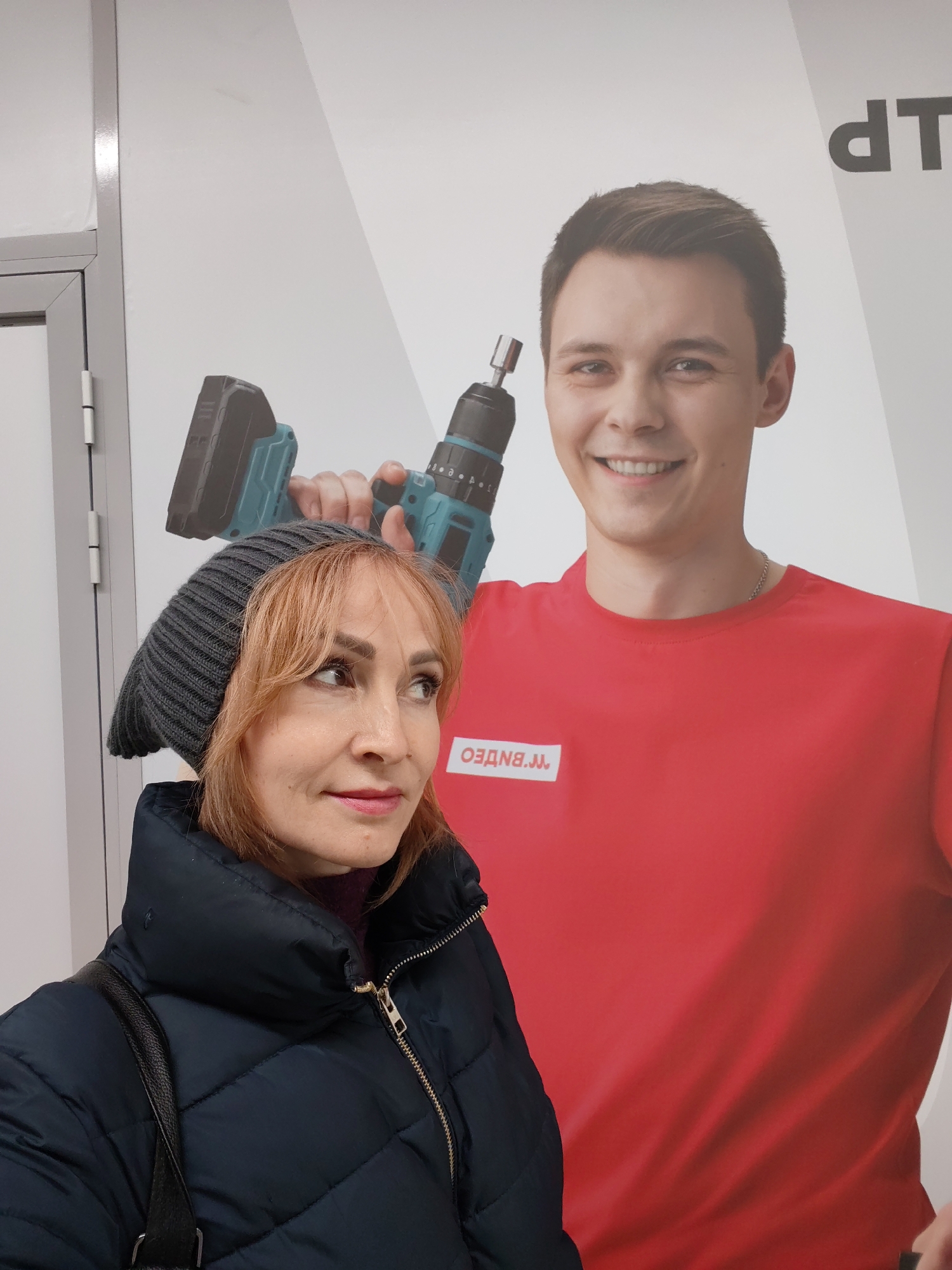 The image depicts a woman standing in an indoor setting, likely a retail environment in Chaikovsky, Russia. In the foreground, a woman with reddish-brown hair and bangs is looking towards her right with a subtle, contemplative smile. She is wearing a dark gray knitted beanie and a dark blue, possibly black, quilted puffer jacket with a high collar. A black bag strap is visible over her left shoulder. Her attire suggests colder weather.
Behind the woman is a large promotional poster featuring a smiling young man. He has short brown hair and is wearing a plain red t-shirt. On the left side of his chest, a white patch displays the reversed text "М.ВИДЕО" (M.VIDEO), indicating a connection to the Russian electronics retailer. The man on the poster holds a blue and black cordless power drill, positioned over his right shoulder, and has a broad, friendly smile.
The background is predominantly the white surface of the poster. To the far left, a gray door frame or architectural column is partially visible, confirming the indoor setting. At the top right corner of the poster, the reversed text "dTP" is partially visible.