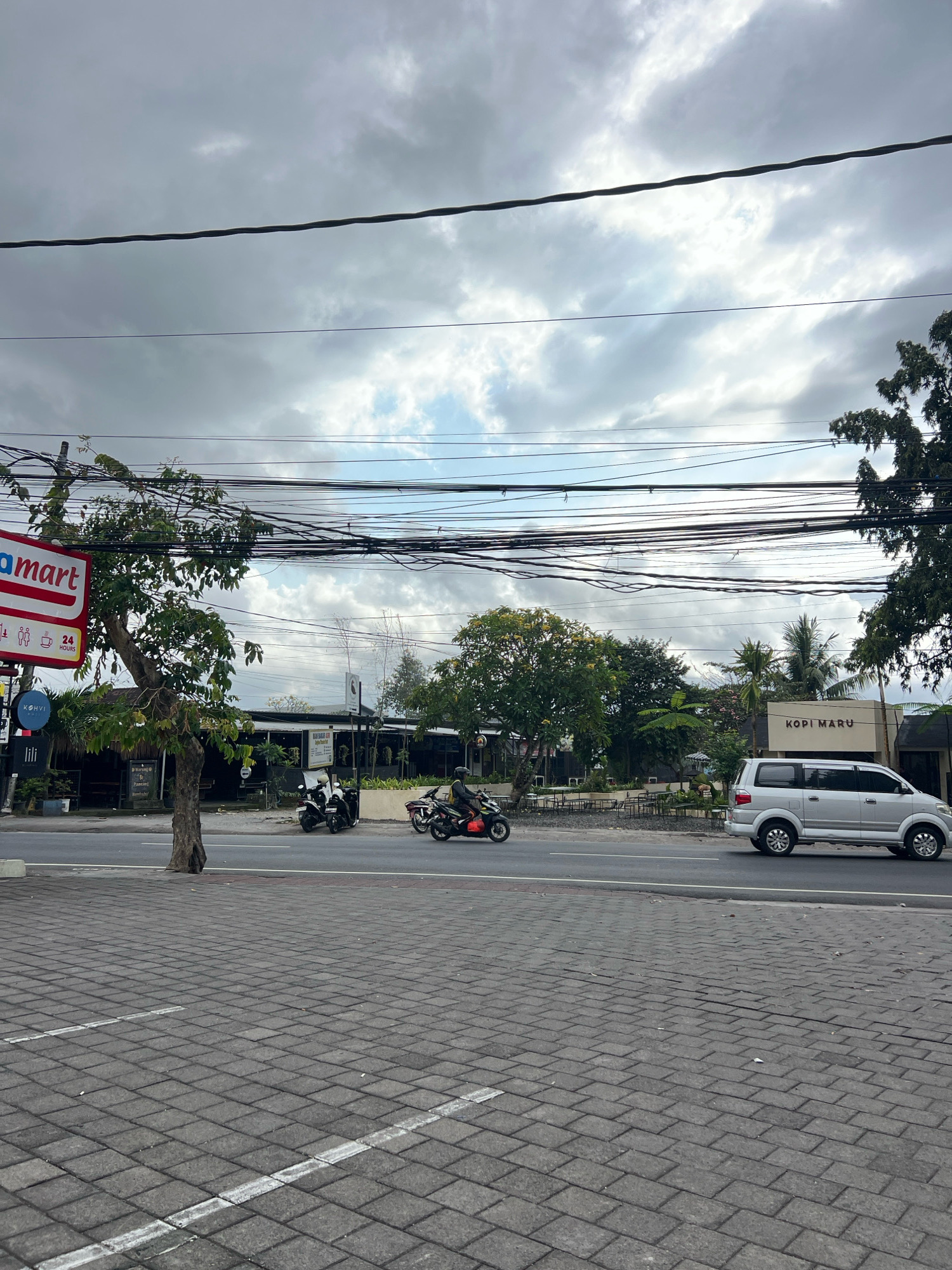 The image captures a street scene in Denpasar, Indonesia, likely near a commercial area given the presence of storefronts and a busy road. In the foreground, a grey paved pedestrian walkway with white painted lines is visible. Beyond the walkway is a road with a silver van and two motorbikes in motion. On the left side of the road, there is a red and white sign for "amart," suggesting a convenience store. Next to it, a small tree with green foliage stands. Further down the road, a building with "KOPI MARU" written on its facade is noticeable, indicating a coffee shop or restaurant. The background shows more trees and buildings under a cloudy sky with visible power lines overhead. The overall scene depicts a typical day in an urban environment with traffic and commercial activity.