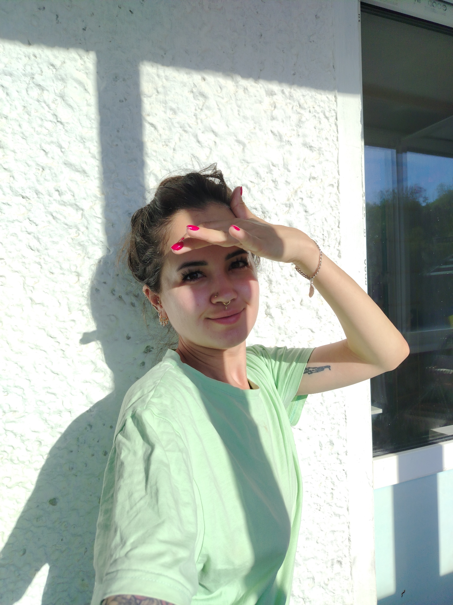 The image shows a young woman, likely in her early twenties, standing outdoors against a textured white wall. She has dark hair pulled up in a bun and is wearing a light green t-shirt. Her right hand is raised to shield her eyes from the sun, revealing bright pink nail polish. She has a nose ring and an earring. A delicate bracelet adorns her right wrist, and a tattoo is visible on her right forearm. The sun casts a distinct shadow of a window frame and horizontal bars on the wall behind her. To her right, a dark-tinted window is visible, reflecting the surrounding trees and sky. The overall setting appears to be an exterior wall of a building in an urban environment, possibly a residential or commercial area in Noviy, Russia.