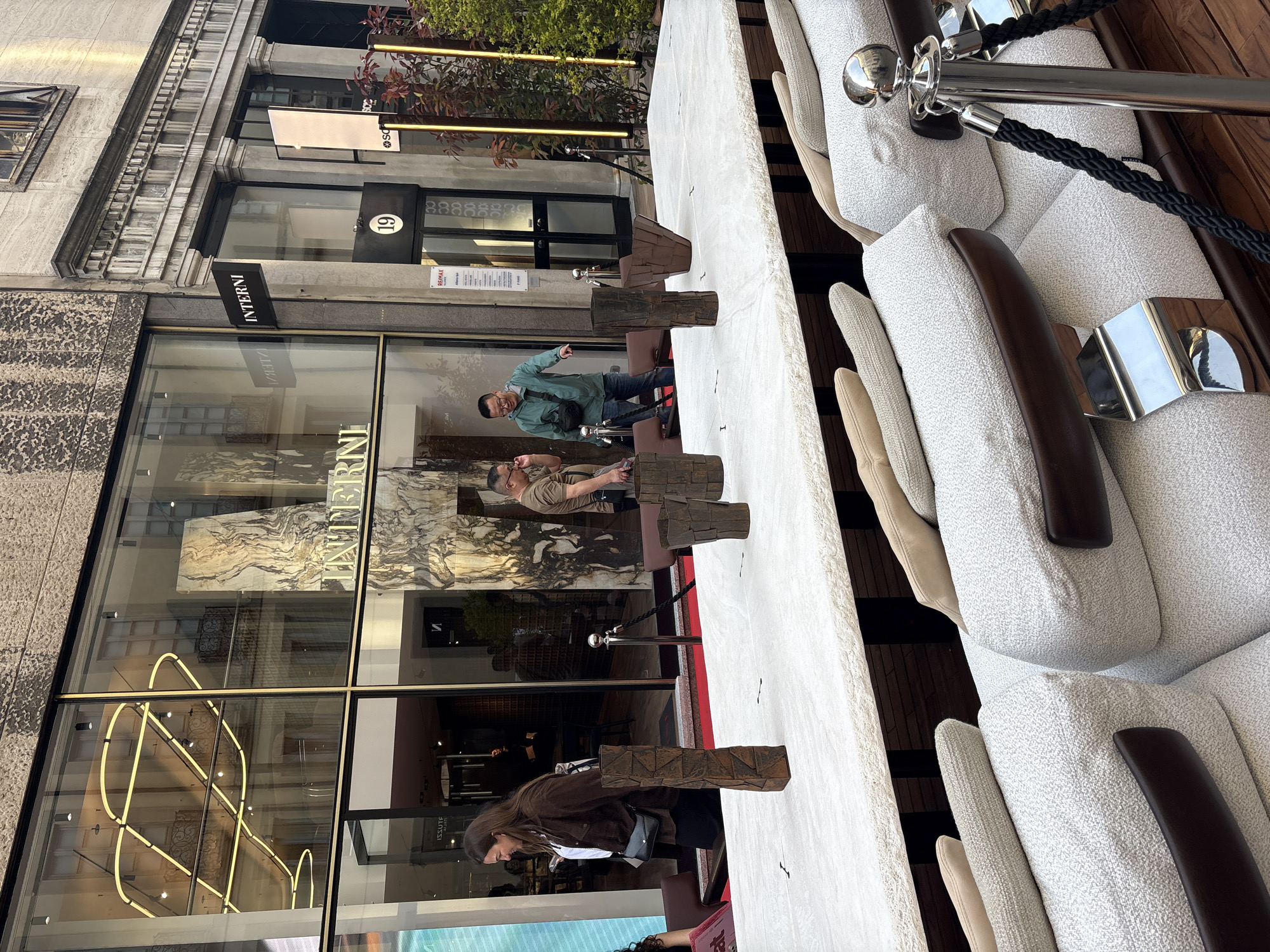 An outdoor scene captures a segment of a bustling street, featuring a modern, elongated seating area situated in front of a building with large display windows. On the right, several light grey, textured lounge chairs with dark wooden armrests are arranged behind a black rope barrier supported by chrome stanchions. A long, light-colored, possibly stone or concrete table runs parallel to the chairs, featuring dark, rustic-looking wooden block decorations.
Three individuals are visible interacting near the table. A man in a light teal jacket, jeans, and glasses points off to the left with his right hand, seemingly indicating something. Another man, wearing a light brown t-shirt and glasses, stands or leans at the table, looking towards the pointing man. A woman with long dark hair, dressed in a brown jacket, stands at the table looking downwards.
The building on the left has a stone facade with intricate architectural details, including cornices. Large display windows reflect parts of the street and a striking interior neon light fixture with an abstract, circular design. Visible text includes "INTERNI" on a black sign above a window and reflected on the glass. The number "19" is displayed above a doorway. A partially visible "RE/MAX" sign can also be seen. The scene is brightly lit by natural daylight, suggesting midday or early afternoon. The overall setting appears sophisticated, indicative of an urban commercial area like Milan, Italy.