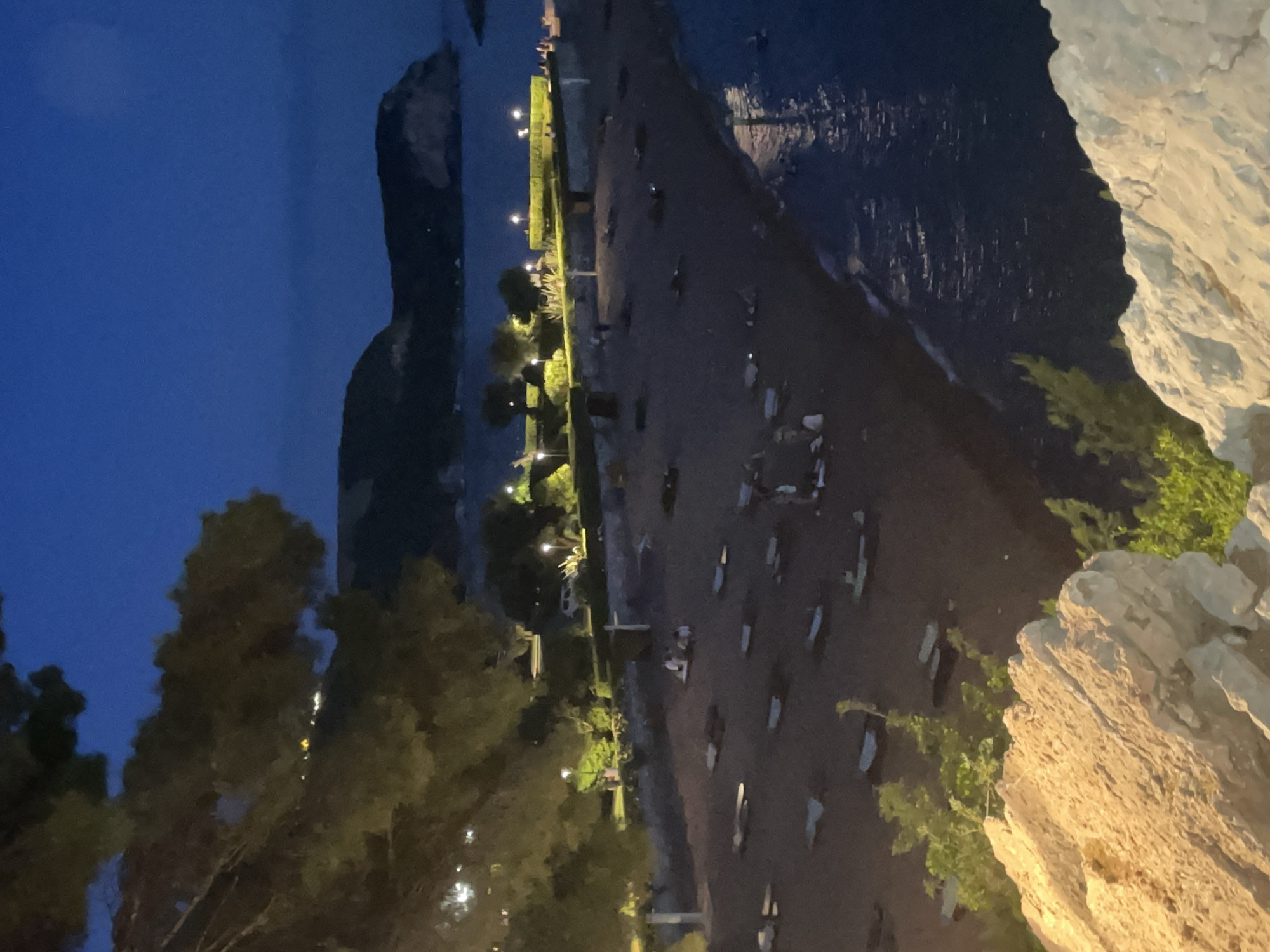 The image depicts a nighttime scene on a beach in Sveti Stefan, Montenegro, viewed from a slightly elevated perspective, likely a rocky outcrop. The sky is a deep twilight blue. In the foreground, large, light-colored rocks with rough textures are visible, with some green foliage growing among them.
Dominating the mid-ground is a sandy beach, sparsely populated with individuals. Several beach chairs or loungers are spread across the sand, some occupied, others empty. A small group of people are gathered together on the beach, appearing to be conversing. Along the edge of the beach, a paved walkway is illuminated by streetlights, with trees and bushes lining the pathway. A white van is parked along the walkway.
The background features the calm sea, with subtle reflections of lights on its surface. Beyond the sea, a dark, hilly landmass, likely an island or a peninsula, is silhouetted against the night sky. There are a few scattered lights visible on this distant landmass. The overall atmosphere is tranquil, with the beach appearing to be winding down for the night.