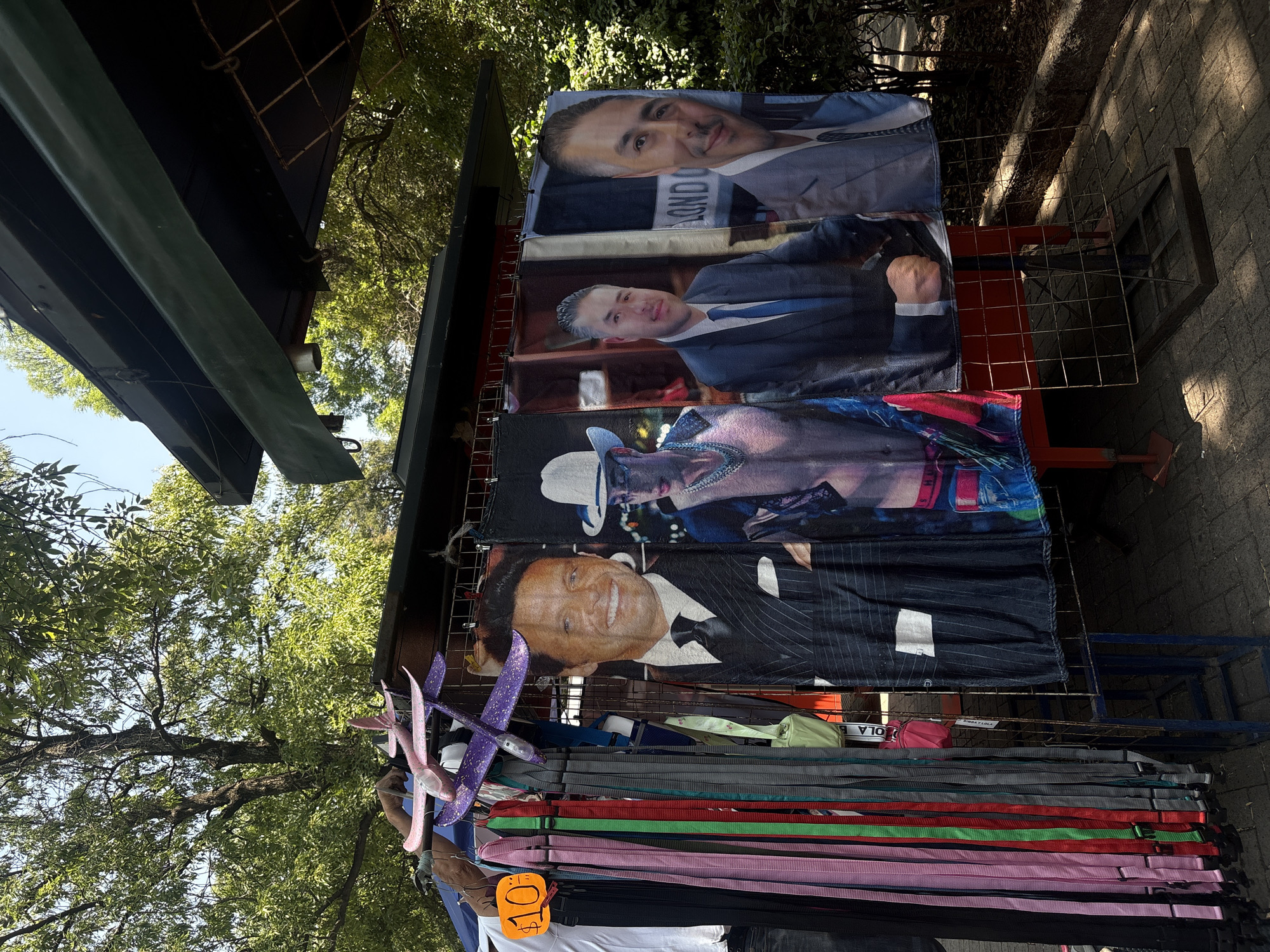 This image depicts an outdoor market stall, likely in Mexico City, under bright daylight. The stall is laden with various merchandise, predominantly large hanging textiles that appear to be blankets or tapestries.
Prominently displayed are four such textiles, each featuring a portrait of a man. The top textile shows a man with short dark hair, wearing a light-colored suit jacket and dark tie. Below him is a younger man with grey-streaked hair, dressed in a dark suit and blue tie, with the word "LONDON" subtly visible on his tie. The third textile features a man in a white cowboy hat and a patterned shirt or jacket. At the bottom, a textile displays the smiling face of the singer Luis Miguel, wearing a pinstripe suit and a black bow tie.
Below these hanging portraits, a stack of colorful straps or belts is arranged, showcasing various hues including red, pink, green, grey, and black. An orange price tag attached to these items clearly shows "10" followed by a dollar sign. To the left, two novelty foam gliders, one purple and one pink, are also on display.
The stall has a dark, possibly green, canopy or roof providing shade. The ground to the right is paved with bricks or pavers. In the background and upper parts of the image, lush green trees and foliage are visible, indicating an outdoor, possibly park-like or tree-lined street setting. The bright sunlight casts distinct shadows, suggesting a clear and sunny day.