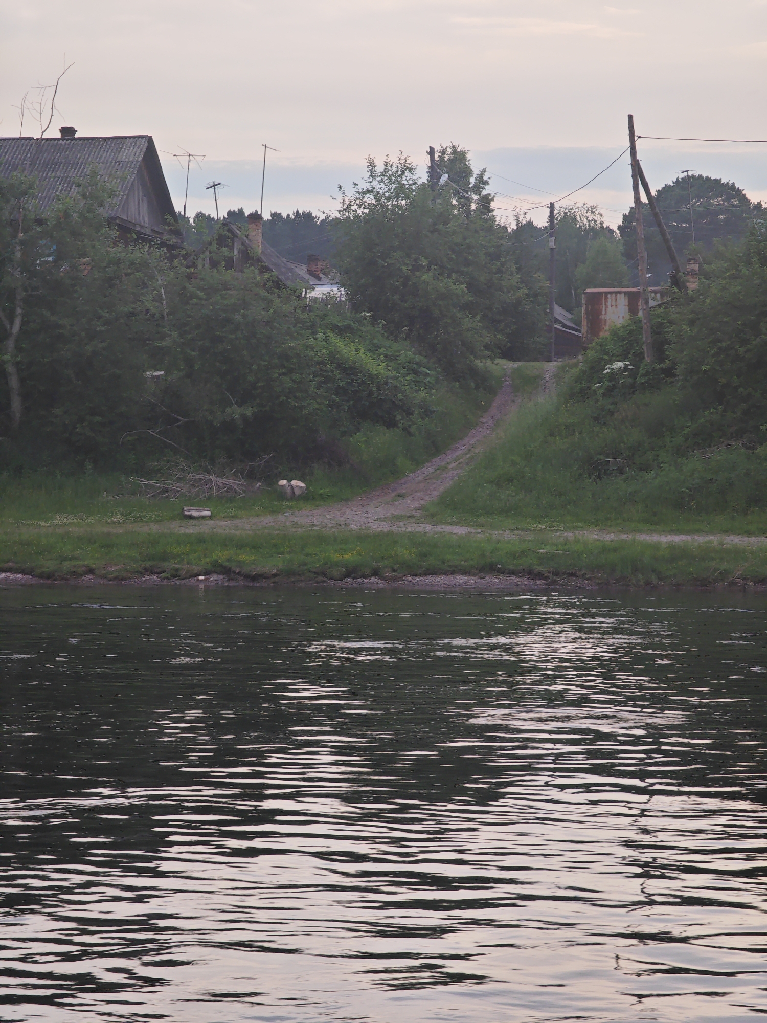 The image depicts a rural scene at dusk, likely in the village of Yukseevo, Russia, characterized by a body of water in the foreground and a slight incline leading to vegetation and buildings in the background. The water exhibits ripples reflecting the muted, overcast sky, which has a pale pink and grey hue.
On the embankment, a dirt path winds uphill, bordered by green grass and dense foliage. Several trees and bushes create a thick natural barrier. Visible on the left side of the path are two roughly cut logs, and a piece of what appears to be a long wooden beam or bench lies on the grass. Further up the slope, a dilapidated wooden house with a corrugated metal roof is partially obscured by trees. Adjacent to it, another structure with a brick chimney is visible. Electrical poles with wires are scattered across the scene, suggesting a connection to a power grid. In the distance, a rusted metal structure, possibly a silo or storage container, stands amidst the greenery. The overall atmosphere is tranquil and somewhat melancholic, indicative of a quiet evening in a rural setting. There are no people or animals clearly visible in the image.