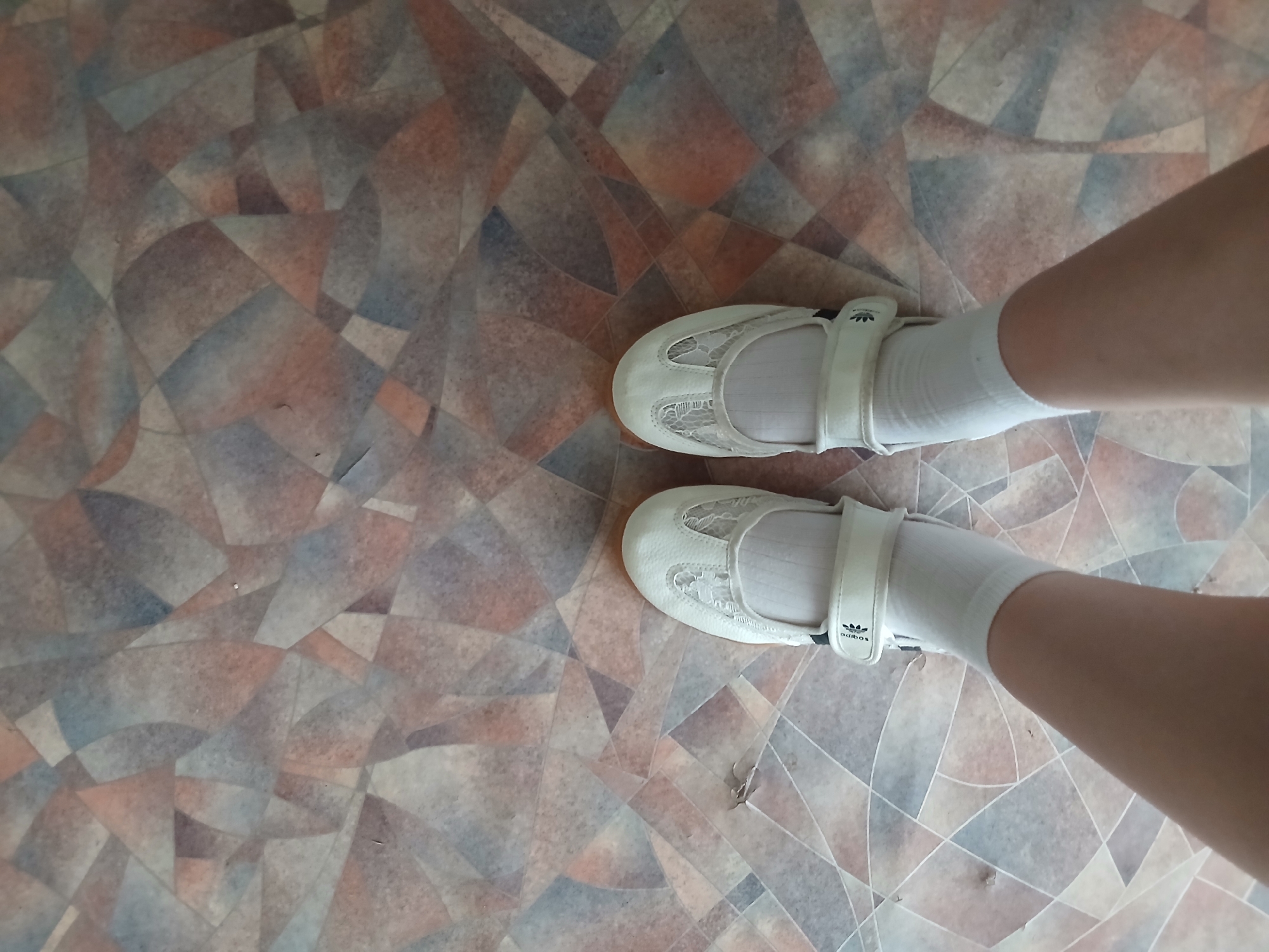 The image is a top-down view of a person's feet wearing white shoes and socks, standing on a patterned floor. The floor has a geometric design with irregular shapes in shades of brown, gray, and blue. The shoes are white, low-cut, with a velcro strap across the instep and lace-like detailing on the vamp. The socks are white and crew-length. A small piece of debris is visible on the floor near the left shoe. The scene is indoors, likely a room in a residential or public building. No specific location in Kum-Aryk, Kyrgyzstan can be determined from the visual information provided.