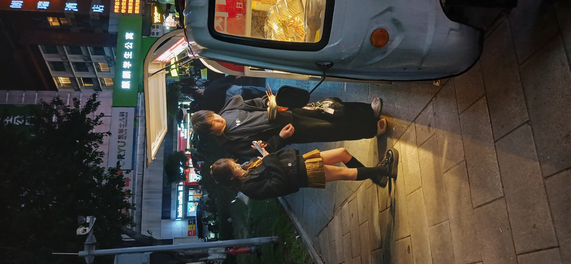 Here is a description of the image:
The image shows a street scene at night in Jiulongpo District, China. Two people, an adult and a younger person, are standing next to a food vendor's cart. The adult is holding food in one hand and possibly a wallet or bag in the other. The younger person is looking at a cell phone. They are standing on a paved sidewalk. In the background are buildings with illuminated signs in Chinese characters. A tree is visible to the left of the frame. A traffic light pole and a fire hydrant are visible at the bottom left of the frame. The signs say: "Lang Rui Hotel", "Student Apartments", and "RYU Pediatrician".