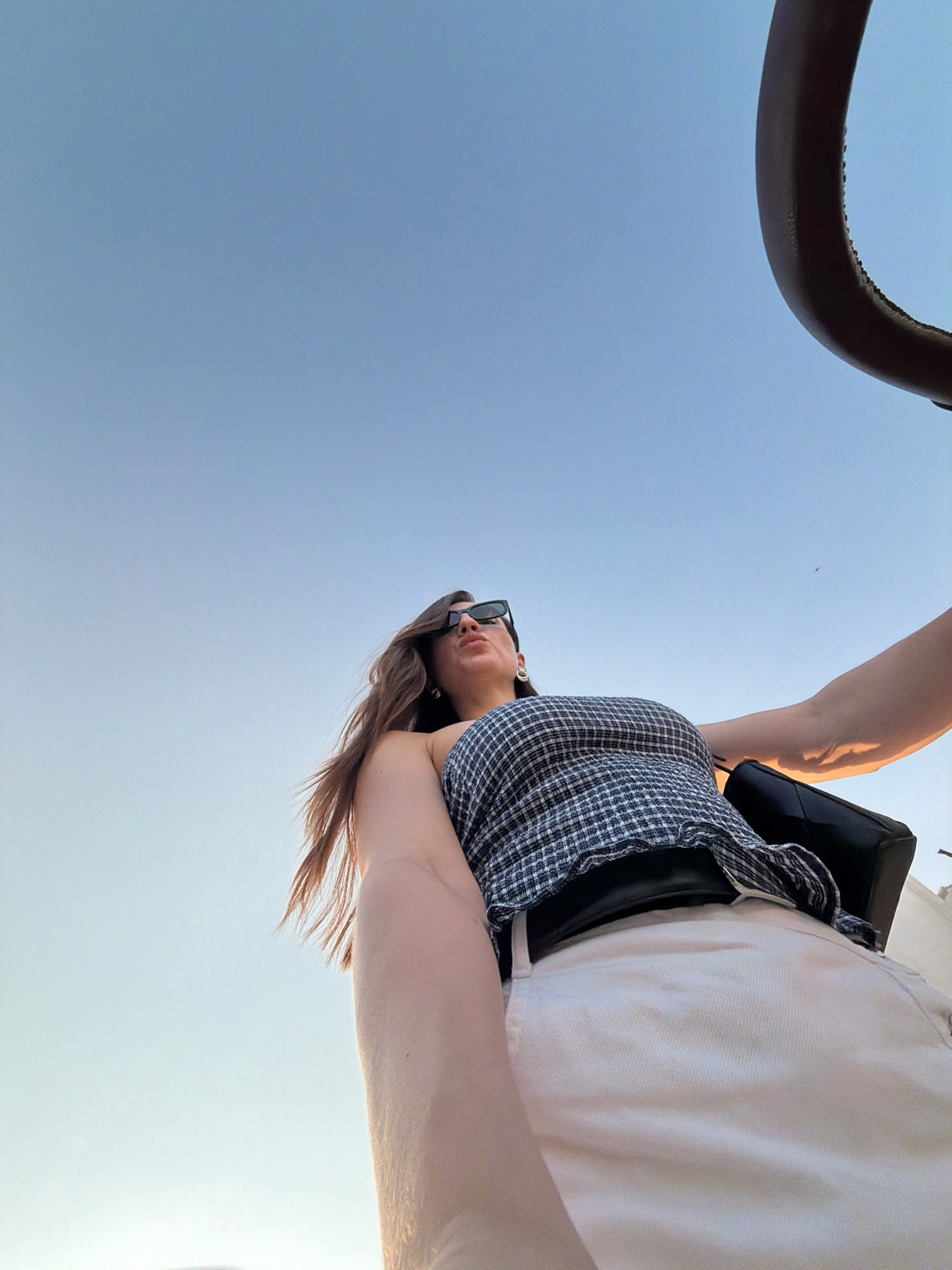 A low-angle, full-body shot from below captures a woman against a clear, pale blue sky, likely at sunset or sunrise, possibly in Sharm El Sheikh, Egypt. The woman is wearing a blue and white checkered crop top, white high-waisted pants, and black sunglasses. Her long brown hair is blowing to her left. Her left arm is extended, and a portion of a dark, possibly leather, handbag is visible at her waist. A dark brown, curved object, which could be part of a railing or a vehicle, frames the top right of the image. The lighting suggests an outdoor setting with diffused sunlight.