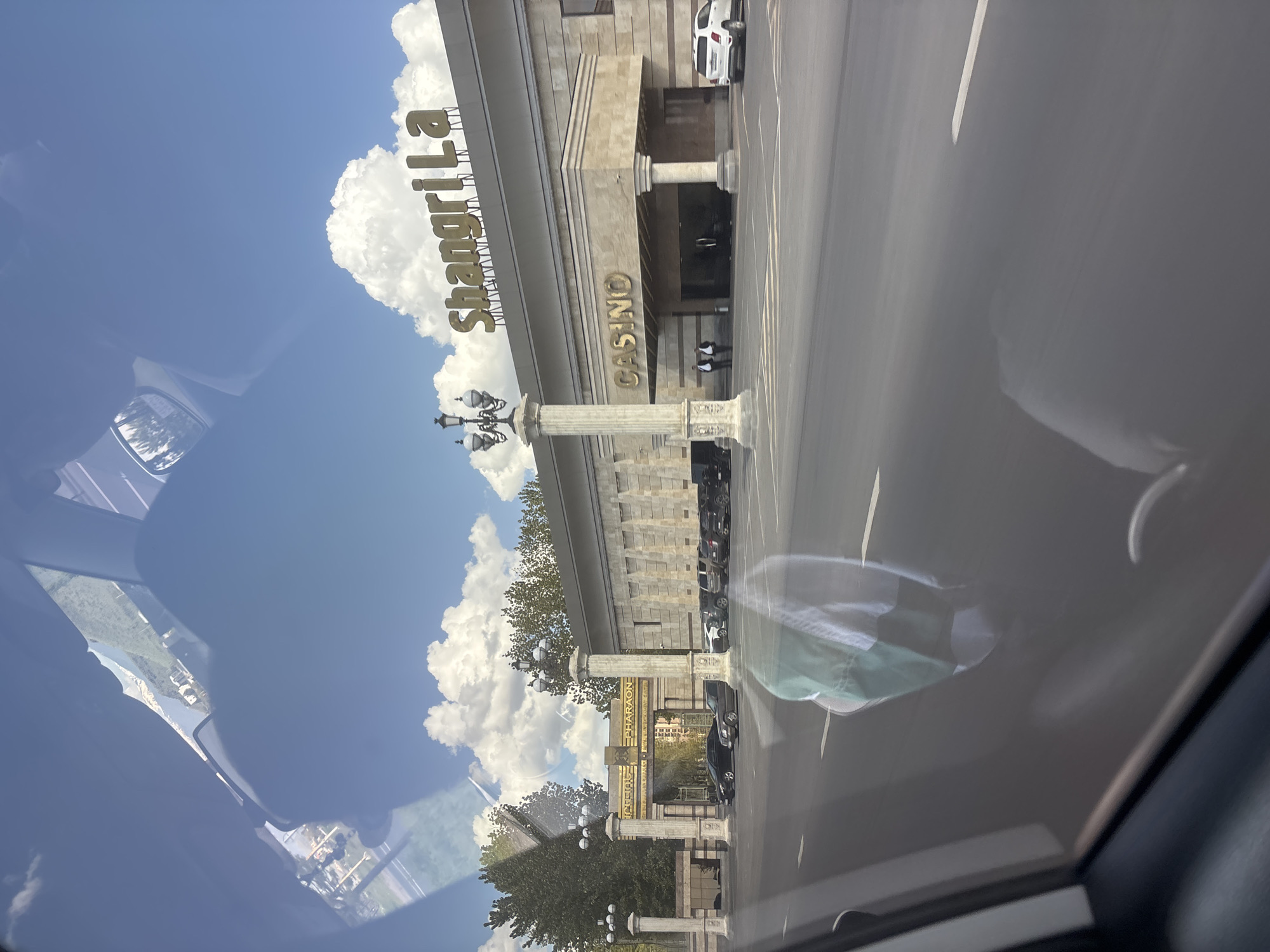 The image depicts a view from inside a car, oriented towards the Shangri La Casino in Verin Ptghni, Armenia. The camera is positioned from the front passenger seat. The casino building is visible in the middle of the frame. Cars are parked in front of the casino. The sky is blue with white clouds. The reflection on the windshield is distracting.