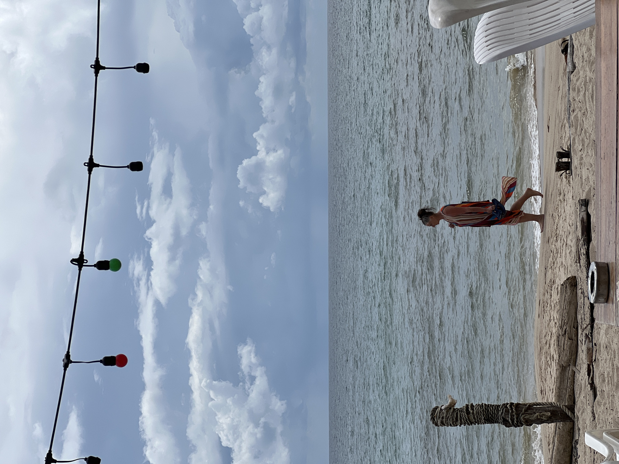 A woman walks barefoot along a sandy beach next to the ocean. She wears a colorful, striped dress and carries a pair of blue sandals. The ocean has moderate waves and is a gray-blue color under a cloudy sky. String lights with red and green bulbs hang in the foreground. There are wooden posts and debris on the beach, and a white sculptural object is visible on the right. The scene appears to be outdoors at a beachside location.