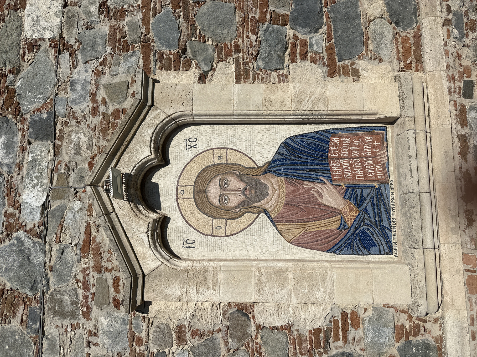 The image shows a mosaic of Jesus Christ on the exterior wall of the Church of Panagia Eleousa in Agros, Cyprus. The mosaic is set within a stone niche above a carved stone ledge. Jesus is depicted with a halo, wearing a blue robe and a reddish-brown tunic. He is holding an open book with inscriptions in Greek. The surrounding wall is constructed of rough-hewn stone and bricks. The inscription at the bottom of the mosaic reads "AWPЄA TΩ OMIΛΩ ΓYNAIKWN ATPΩ".