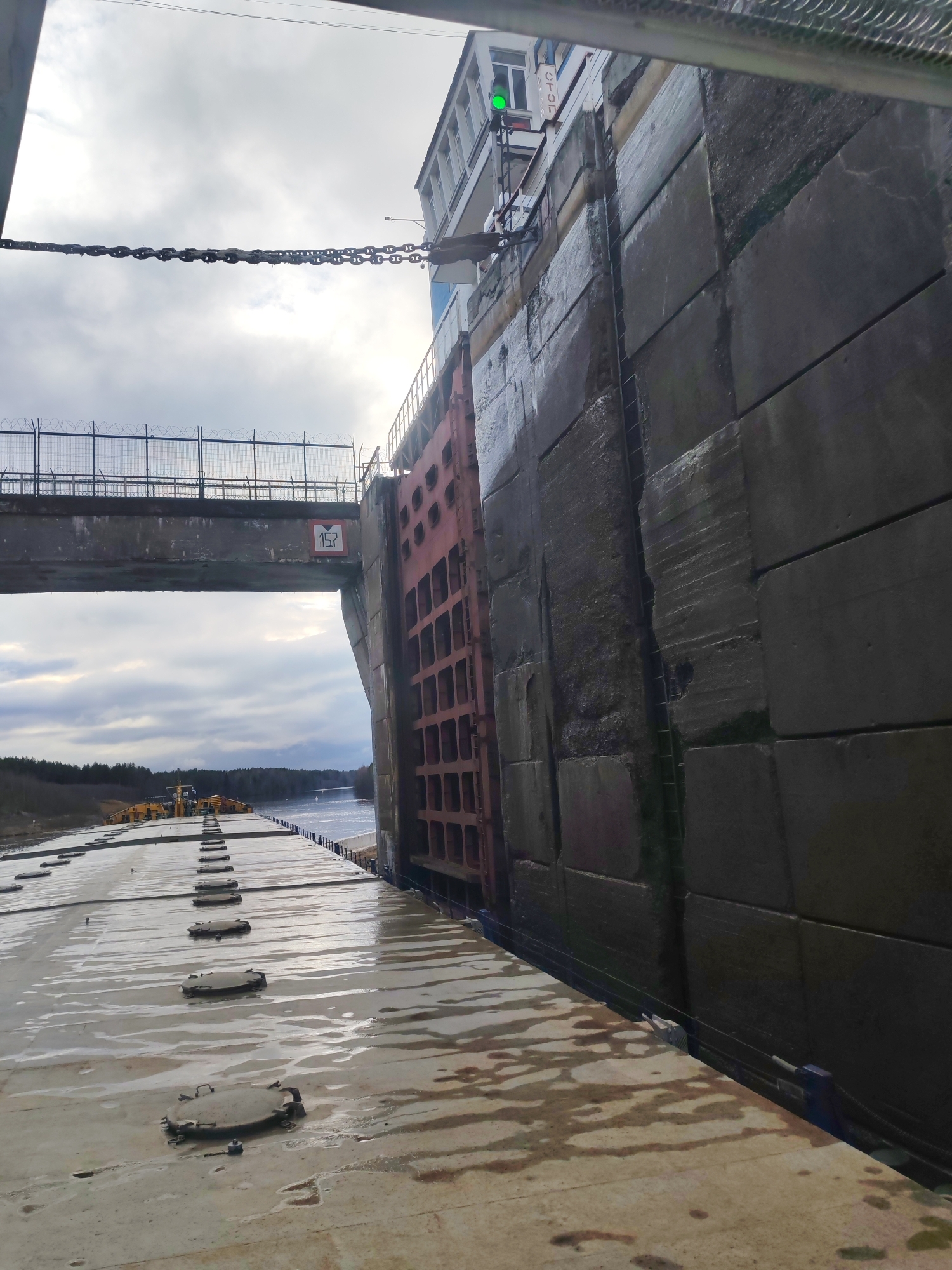 The image shows a large, possibly industrial, waterway lock located in an unknown city. A massive concrete wall is prominently featured, marked with visible seams between its block-like sections. A red metal gate, presumably part of the lock system, is adjacent to the wall. A metal bridge-like structure spans the waterway, with a sign displaying the number "157" attached to its side. The deck of a vessel is visible in the foreground, its surface wet and dotted with access hatches. In the background, a narrow body of water stretches into the distance, flanked by a treeline. The sky above is overcast, with diffused sunlight filtering through the clouds. A chain connects the lock wall to a nearby building.