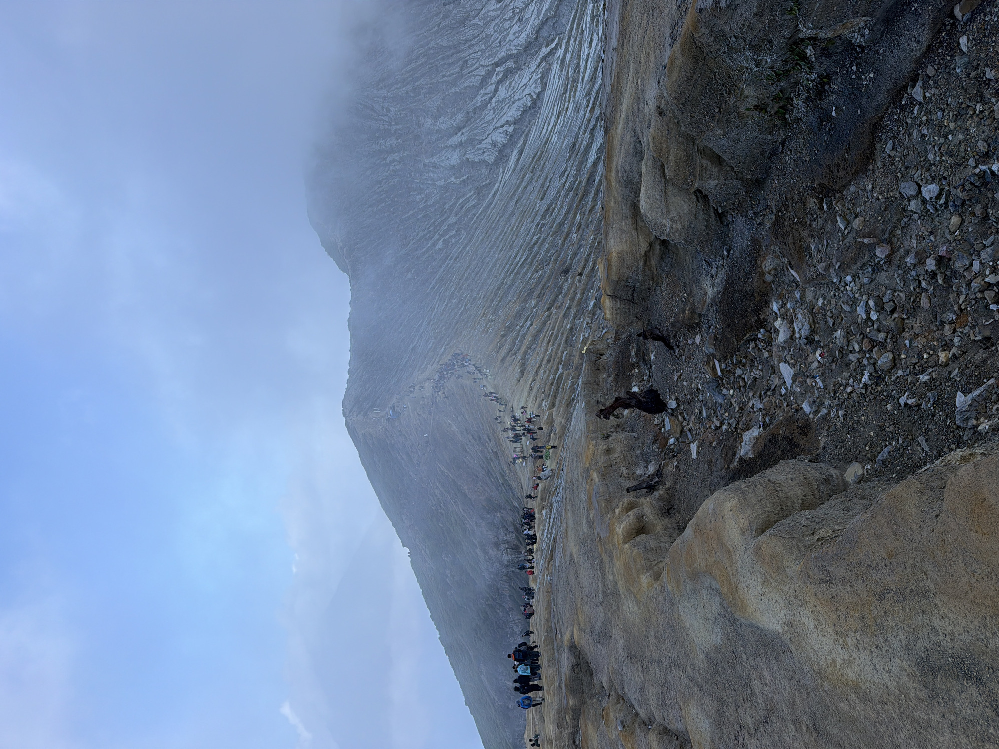 The image depicts a volcanic landscape with a large group of tourists ascending a ridge. The terrain is rocky and barren, with a path carved into the side of a mountain. The sky is overcast with a pale blue hue. The tourists, dressed in casual clothing, are walking in single file up the steep incline. Some are carrying backpacks, and others are using walking sticks. The mountain itself appears to be made of ash and rock, with evidence of past volcanic activity. The overall scene suggests a popular tourist destination in a remote, mountainous region.