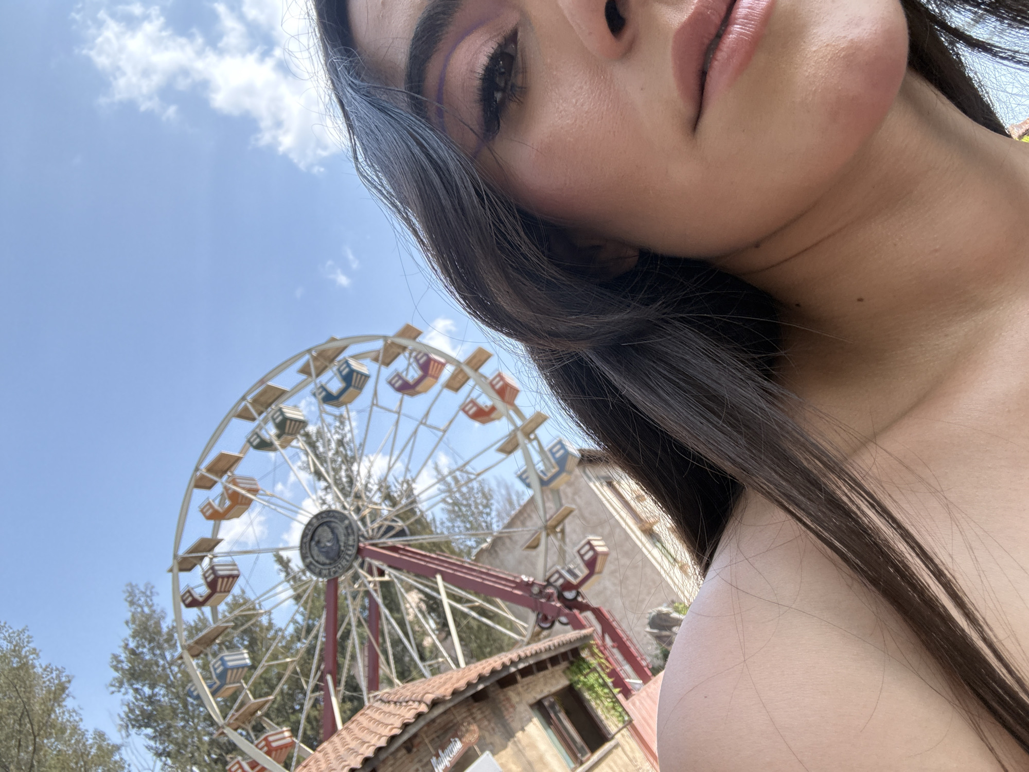The image is a low-angle selfie of a fair-skinned person, cropped from the top of the head to the shoulders. The person has dark hair and is wearing makeup. The person’s face is angled toward the upper right, so that a portion of their forehead, eye, nose, lips, jaw, and neck are visible.
Behind the person, and slightly off to the left, is a colorful Ferris wheel. The Ferris wheel is situated in the city of Domingo Arenas, Mexico and is partially obscured by trees and a building with a red-tiled roof. Above the Ferris wheel, the sky is blue with a few white clouds.
