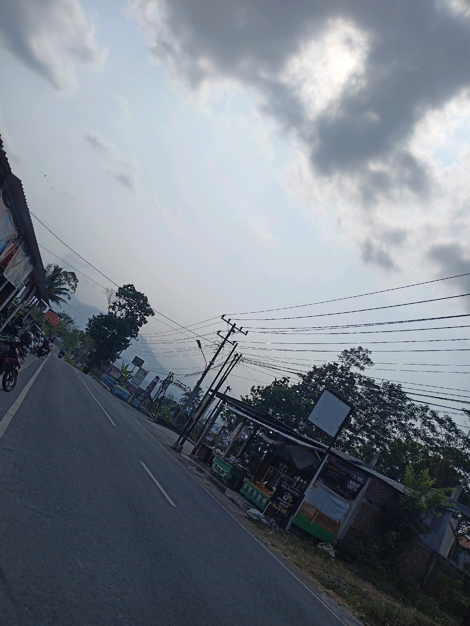The image depicts a street scene in Payaman, Indonesia, characterized by a road angled diagonally from the lower left to the upper right. Along the left side of the road, there are stalls or shops with awnings, and several parked motorcycles. On the right side of the road, there is a row of smaller structures that appear to be businesses, including a stall with green containers and signage, and a building constructed with brick and corrugated metal. Utility poles with numerous wires are prominent in the mid-ground and extend into the background. Trees are visible on both sides of the road, with a larger tree on the right and palm trees on the left. In the distance, a faint outline of mountains is visible against a cloudy sky. The lighting suggests it is either early morning or late afternoon.