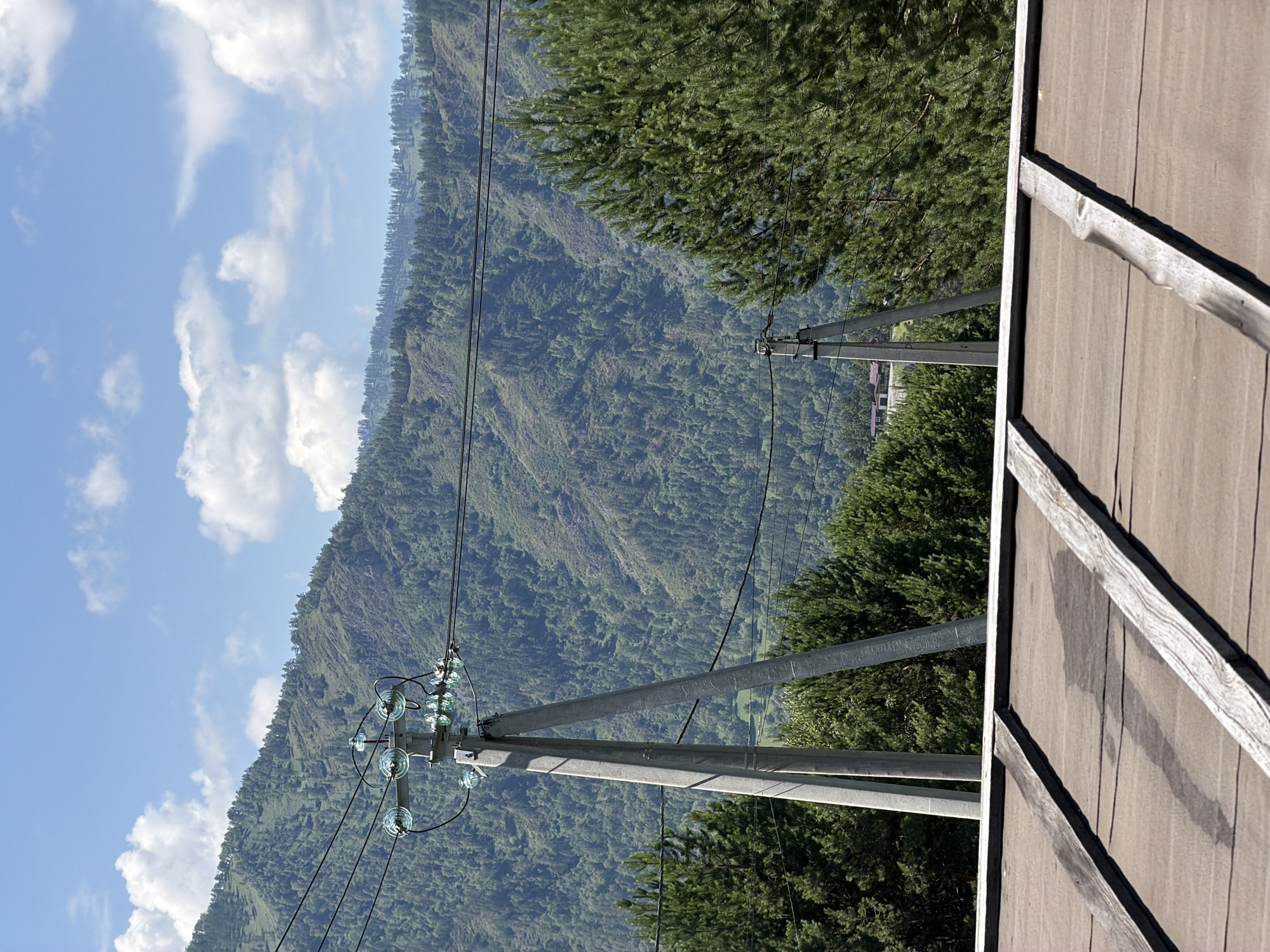 The image depicts a mountainous landscape under a partly cloudy blue sky. In the foreground, the top of a brown roof with wooden beams is visible. Behind the roof, a tall concrete utility pole with glass insulators and electrical wires stands prominently. Further back, a dense forest covers the slopes of a large mountain. The scene is tranquil and natural, suggesting a rural or remote location. A small building is visible in the distance, nestled among the trees. This scene is located in the village of Elekmonar, Russia.