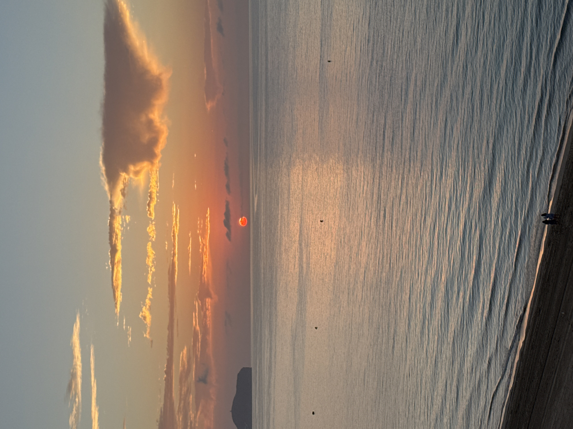 The image captures a serene sunset over the Mediterranean Sea, likely from the coast of Calp, Spain. The sun, a vibrant orange orb, is partially obscured by a thin layer of clouds, casting a warm glow across the sky. The sky transitions from a pale blue at the top to soft oranges and pinks near the horizon, with elongated, wispy clouds catching the last rays of sunlight. In the distance, to the left, a dark, hilly landmass, possibly the Penyal d'Ifac, juts out into the sea. The sea itself is calm, with gentle waves lapping at a sandy beach in the foreground. The water reflects the colors of the sky, creating a shimmering effect. A few dark buoys or markers are visible scattered across the water. On the beach, two small figures, appearing to be people, are walking near the water's edge. The overall scene evokes a sense of tranquility and natural beauty.
