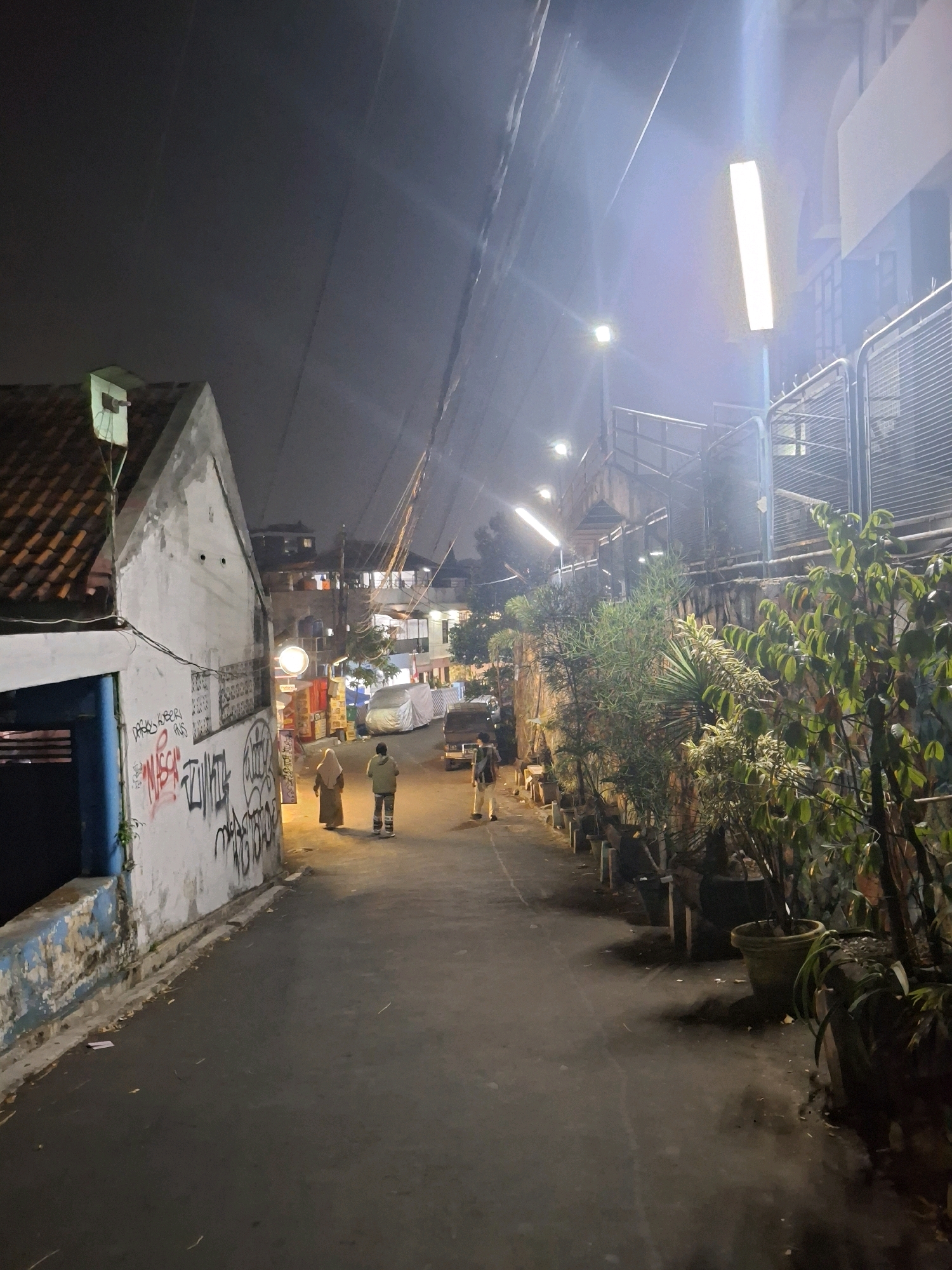 This image depicts a dimly lit street scene in Bandung City, Indonesia, at night. The street is paved with asphalt and appears narrow. On the left side of the street, there is a white building with a red-tiled roof and blue trim. The building's wall is covered in graffiti. Further down the street, other buildings and structures are visible, some with lights on.
Two individuals are walking away from the camera in the middle of the street. One is wearing a light-colored hijab and a dark garment, while the other is wearing a light-colored hooded jacket and pants. Another person, wearing a backpack and light-colored pants, is walking towards the camera in the distance. Several potted plants are arranged along the right side of the street, and a fence or railing is present above them. A bright, vertical street lamp illuminates a portion of the scene. Power lines are visible overhead. The overall atmosphere is urban and somewhat gritty, characteristic of a backstreet in a densely populated city at night.