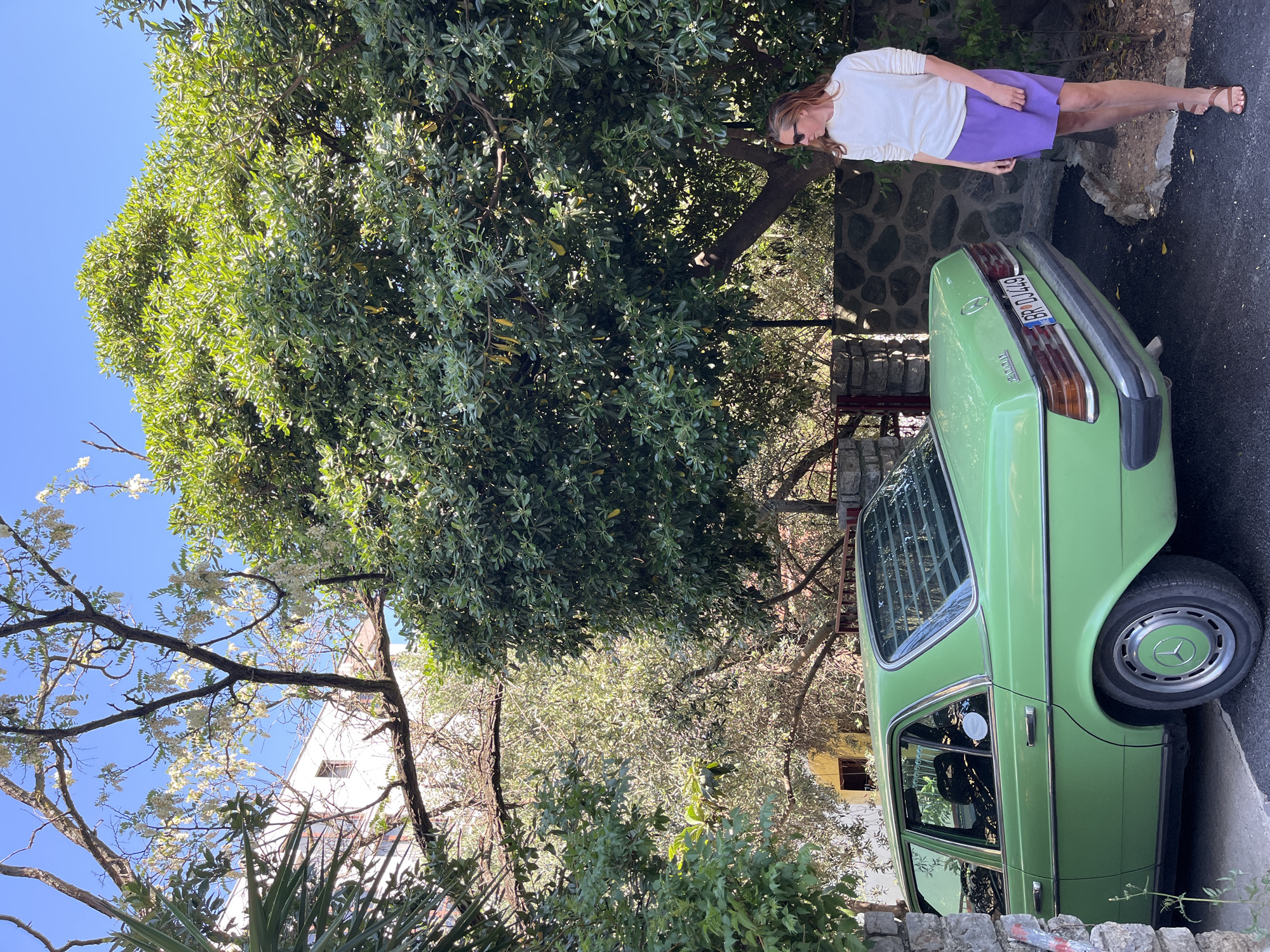 A light green vintage Mercedes-Benz car is parked on a paved surface next to a stone wall in Budva, Montenegro. The car's license plate reads "BD DJ 449." A woman with long hair is standing near the back of the car, wearing a white long-sleeved shirt and a short purple skirt. She is looking downwards. A large, leafy tree casts a shadow over the car and the woman. A white building is visible in the background. The sky is clear blue.