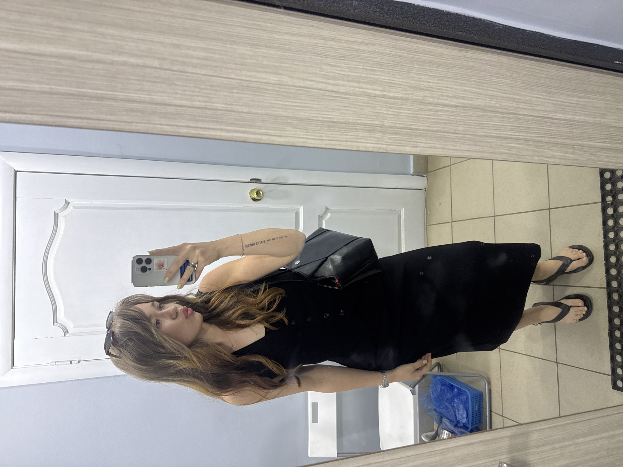 A young woman is taking a selfie in a mirror. She has light brown hair and is wearing a black dress, black flip-flops, sunglasses on her head, and a black purse. She is standing in what appears to be a hallway or a room with a tiled floor and a white door. The lighting is artificial. The location is specified as Chaikovsky, Russia.