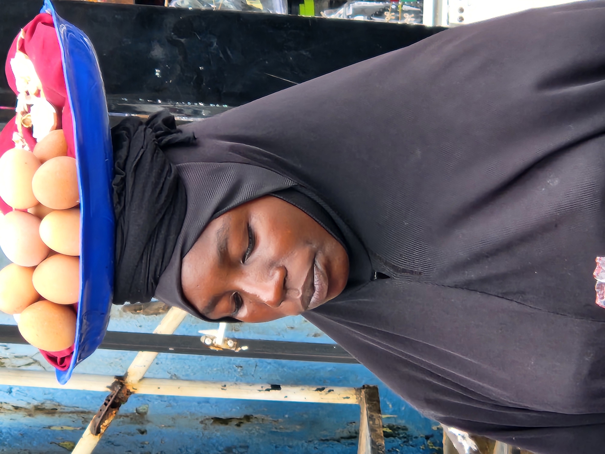 A woman wearing a black hijab and black attire balances a blue tray of eggs on her head in an outdoor market setting in an unknown city. The tray contains approximately six to eight light brown eggs. Behind the woman, a blue, weathered wall with metal bars is visible. To the right of the woman, a dark, reflective surface, possibly a shop window or display, is present, with some colorful items and lights in the background. The woman's face is partially visible, and she appears to be looking down. The overall scene depicts a vendor in a market.