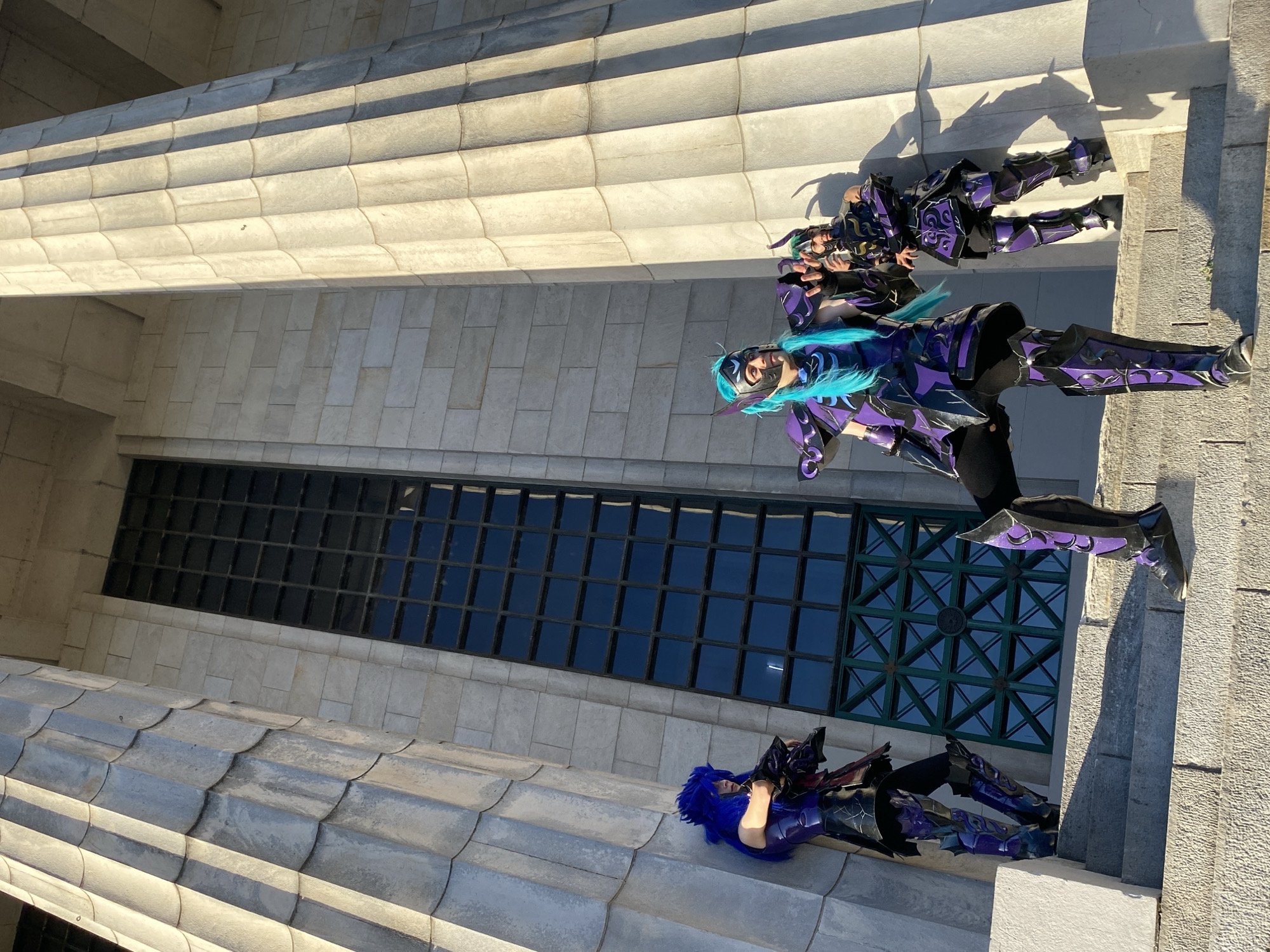 The image shows three people cosplaying in front of a large windowed structure. The three are in detailed, colorful costumes of purple, black, and blue. The person in the center of the frame has a blue wig and is facing the viewer. One person is positioned above the center person, and another below. The background consists of a large window with a grid pattern and light-colored stone. The stone structure and window architecture resembles that of the Law Faculty of the University of Buenos Aires, Argentina.