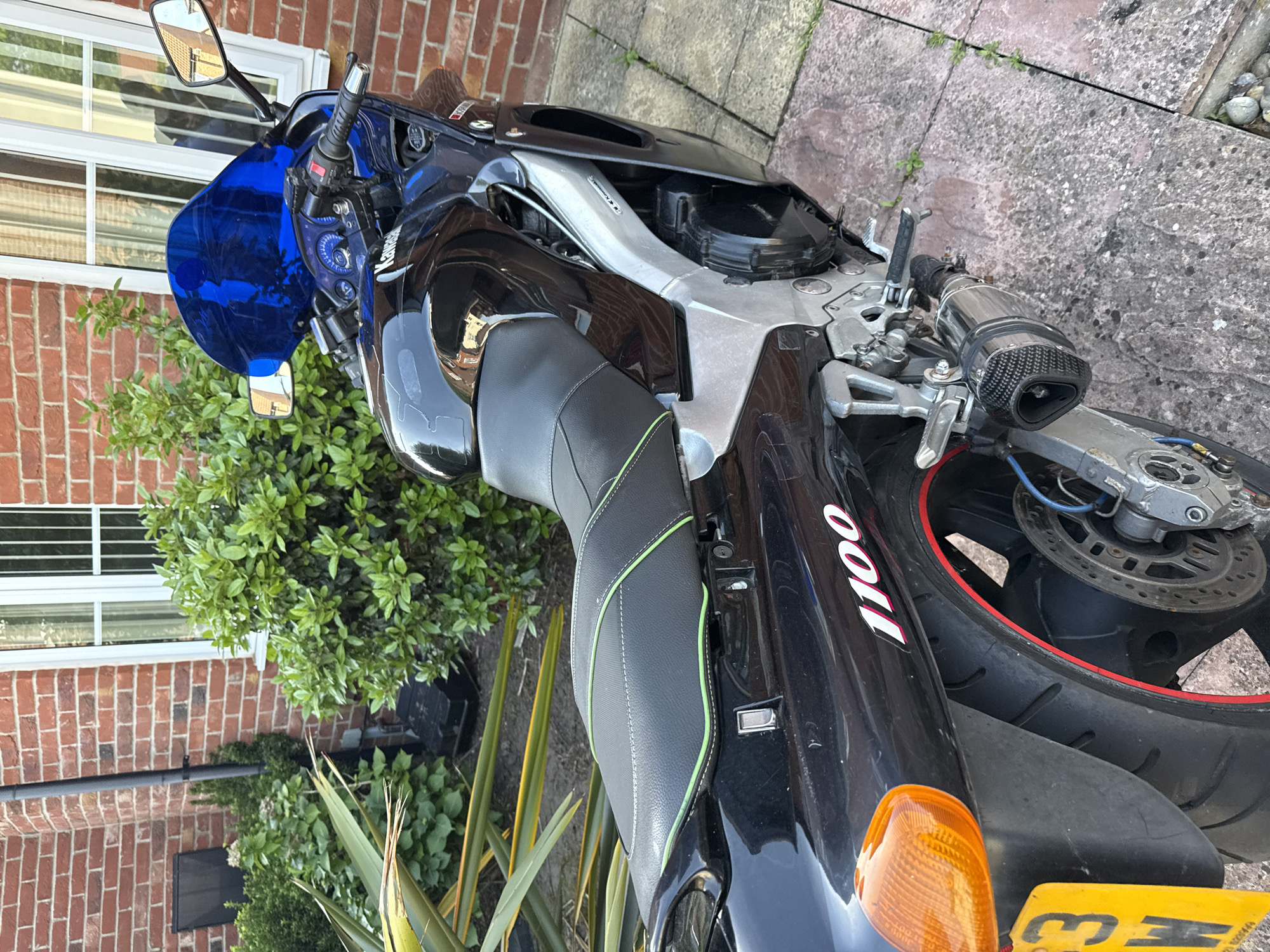 The image shows a black motorcycle parked on a paved area next to a brick building. The motorcycle has a blue windscreen and a black seat with green stitching. The word "1100" is visible on the rear of the bike. A UK license plate is also visible. The surroundings include plants, a brick wall, and a window. The location is Great Yarmouth, United Kingdom, as indicated by the license plate.