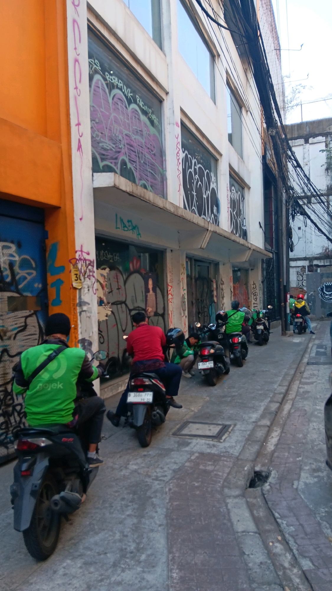 This image depicts a street scene in South Jakarta, Indonesia, characterized by urban decay and commercial activity. The foreground is dominated by several motorcycle riders wearing green vests, indicative of ride-sharing services like Gojek. The buildings lining the street are adorned with extensive graffiti, suggesting a vibrant but potentially unmanaged urban environment. Some buildings feature large glass windows that reflect the surrounding urban landscape and the graffiti. The street is paved with cobblestones and concrete, with a visible drain on the right side. The sky is overcast, casting a diffused light on the scene. The presence of multiple motorbikes suggests a common mode of transportation and economic activity within this area. The overall atmosphere is one of bustling, gritty urban life.