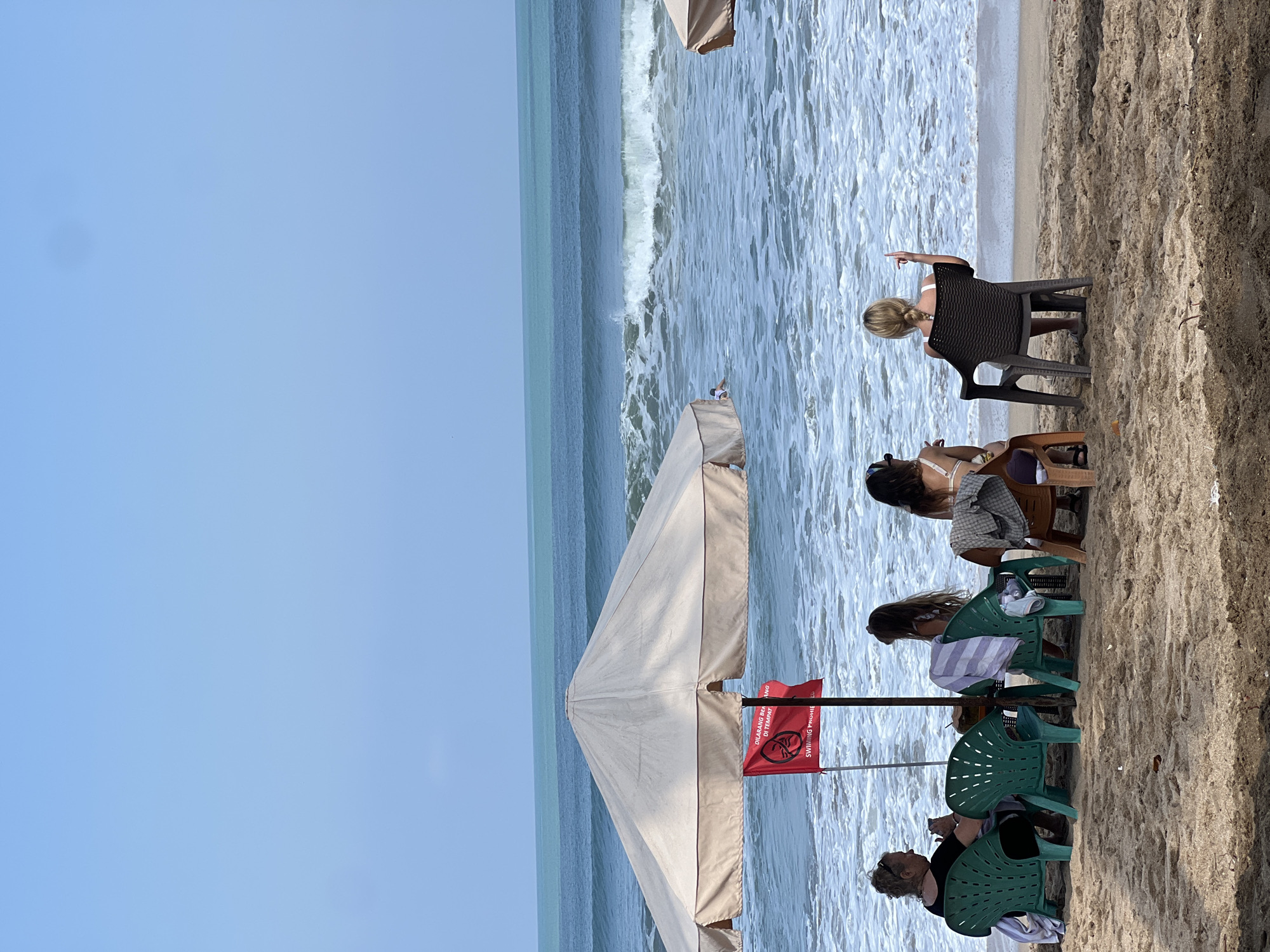 The image depicts a beach scene in Kuta, Indonesia. The foreground shows a sandy beach with several beach chairs set up. Four women are seated in these chairs, facing the ocean. An umbrella is erected over some of the chairs, and a red flag with a prohibition symbol and text is attached to its pole. The ocean in the background has moderate waves breaking on the shore. The sky is clear and blue. One of the women is gesturing with her finger. The overall scene suggests a relaxed atmosphere on a tropical beach.