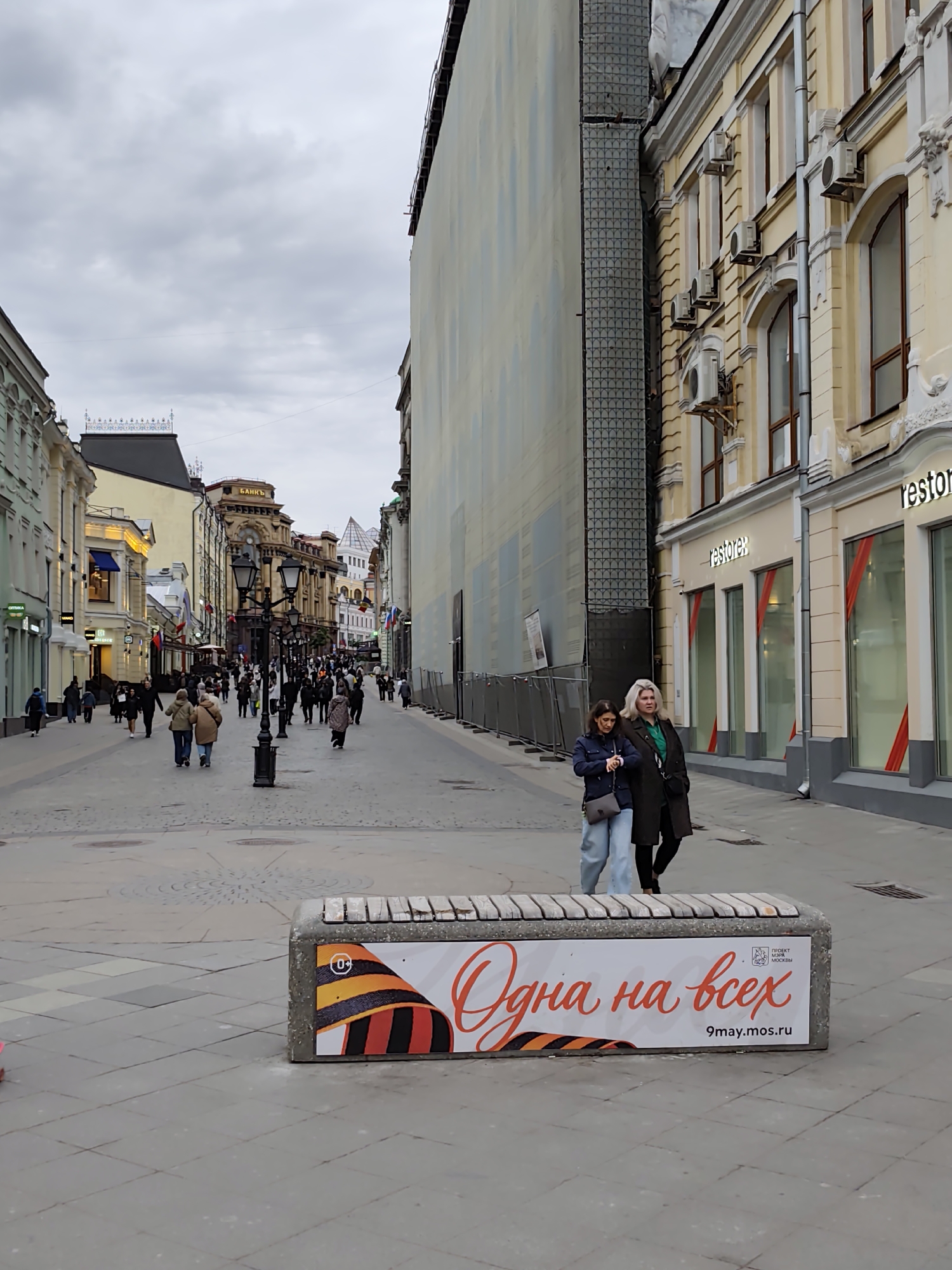 The image depicts a street scene in Moscow, Russia. In the foreground, a concrete bench features an advertisement with a prominent ribbon graphic and the text "Одна на всех" (One for all) followed by "9may.mos.ru". Two women are walking on the sidewalk next to the bench. One is wearing a dark jacket and light blue jeans and the other has blonde hair and is wearing a long dark coat over a green top. The street stretches into the background, lined with buildings of varying heights and architectural styles. A large number of pedestrians are walking in the distance. Street lamps are visible along the street. The sky is overcast. On the right side of the image, the facade of a building is visible, including the sign "restorex" above storefront windows.
