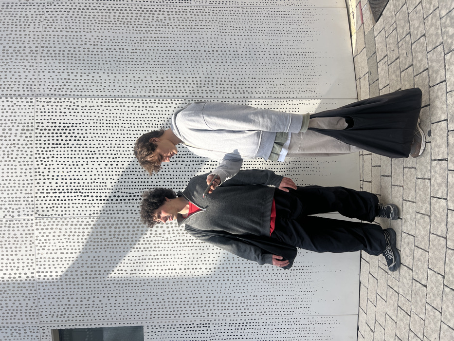 Two males are depicted standing on a paved surface against a white perforated building facade, identified as the Peres Center for Peace and Innovation in Jaffa, Tel-Aviv, Israel.
The male on the left, with dark, curly hair, wears a dark grey quarter-zip sweater over a red t-shirt, black loose-fitting cargo pants, and black athletic shoes. He carries a black fabric shoulder bag on his left side and is observed looking towards his left.
The male on the right, with brown, wavy hair, wears a light grey zippered hoodie layered over a light green and a light beige shirt, light beige baggy pants, and brown athletic shoes. He holds a dark-colored smartphone horizontally with both hands, directing his gaze downward at the screen. He also carries a large black fabric tote bag in his right hand.
The background facade features a pattern of vertical lines composed of circular perforations. A dark rectangular opening is visible on the far left of the facade. The ground is paved with light-toned rectangular blocks. The scene is illuminated by bright direct sunlight, casting distinct shadows.
