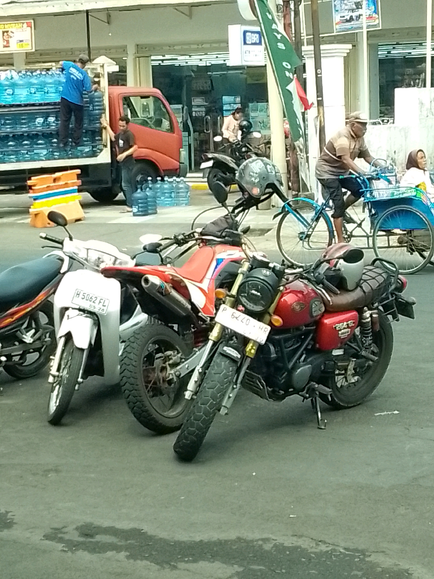 In Jember, Indonesia, on a street, a scene unfolds with a red truck laden with bottled water, suggesting a delivery. Two men are engaged in the loading or unloading process. One man, wearing a blue shirt, stands in the truck bed, handling the water bottles. Another man, in a dark shirt, stands beside the truck. Several stacks of colorful plastic crates are visible near the truck, likely used for transporting goods. In the background, a BRI ATM is visible, indicating a commercial area. The street is populated by several motorcycles and a bicycle rickshaw. An elderly man rides the rickshaw, accompanied by a woman. A green flag hangs from a pole, bearing Indonesian text. The ground appears to be wet in patches, possibly from rain or recent cleaning. The overall impression is one of everyday commerce and transportation in a bustling Indonesian city.