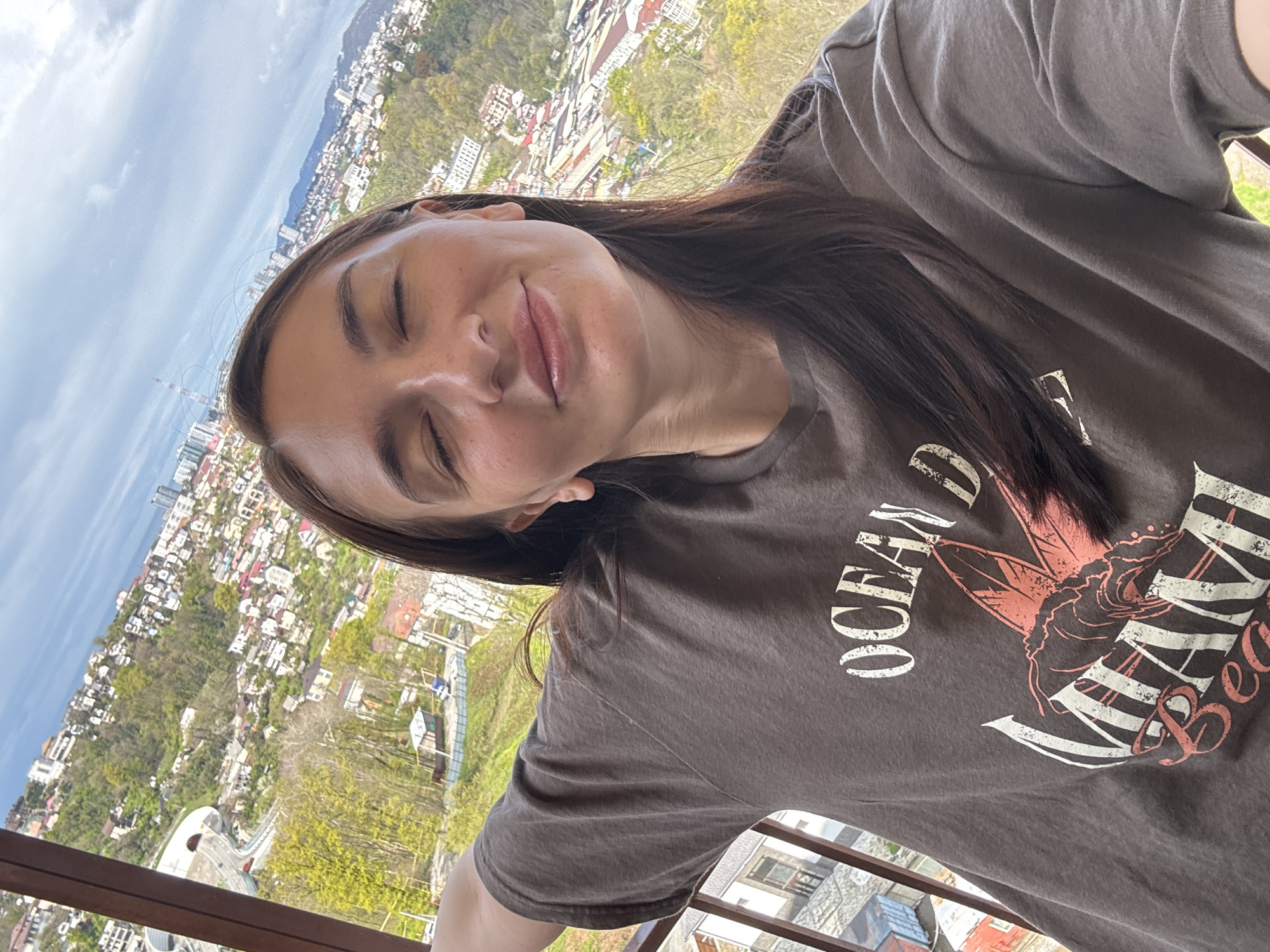A selfie of a woman with closed eyes, taken at an angle on what appears to be a balcony. She has long, dark hair and is wearing a gray T-shirt with the words "OCEAN MIAMI BEACH" on it. The background reveals a cityscape with buildings, trees, and hills. The location is Sochi, Russia.