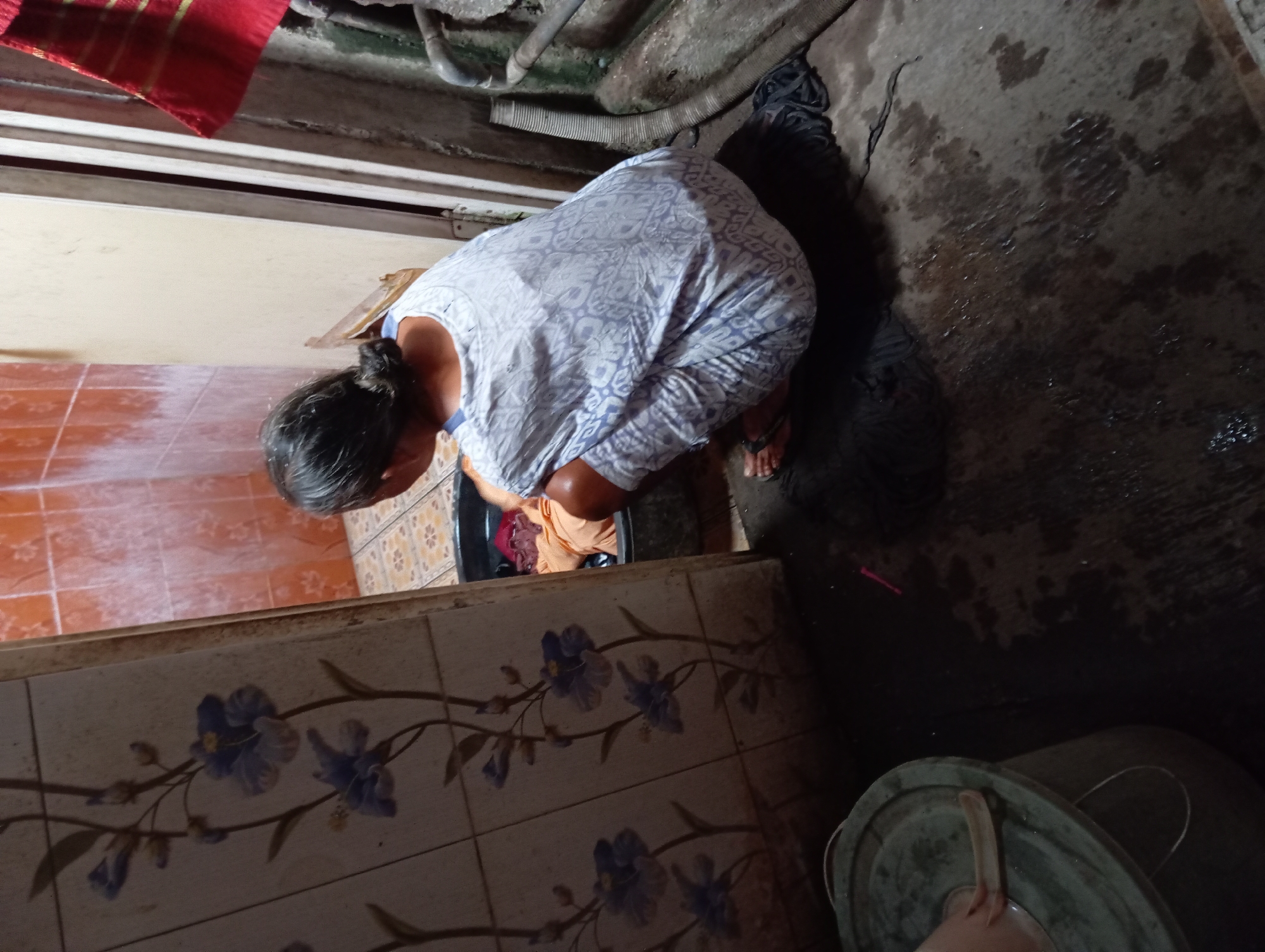 A person with graying hair pulled back in a bun is hunched over a black basin, seemingly washing clothes in a tiled bathroom. The person is wearing a white and blue patterned top and orange bottoms. The bathroom features orange tile walls on one side, and floral patterned tiles on the other side. Above, exposed pipes run across the ceiling. The floor is wet, and a dark gray bucket with a green lid sits in the lower right corner. Based on the location provided, the image appears to be in a residential bathroom in the city of Baron, Indonesia.